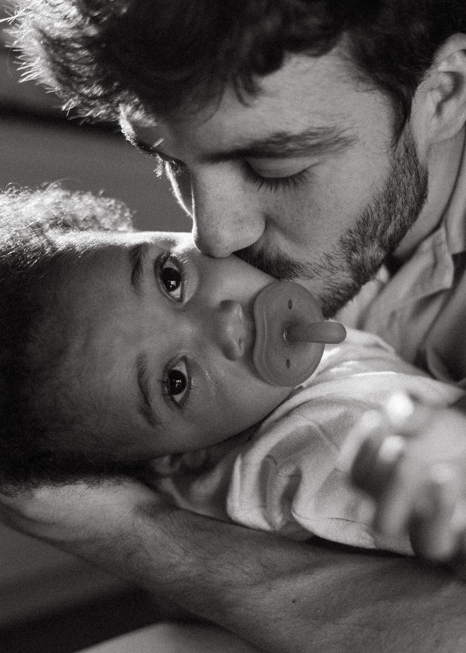 Black and white photo of a bearded father kissing his pacifier-holding newborn baby.