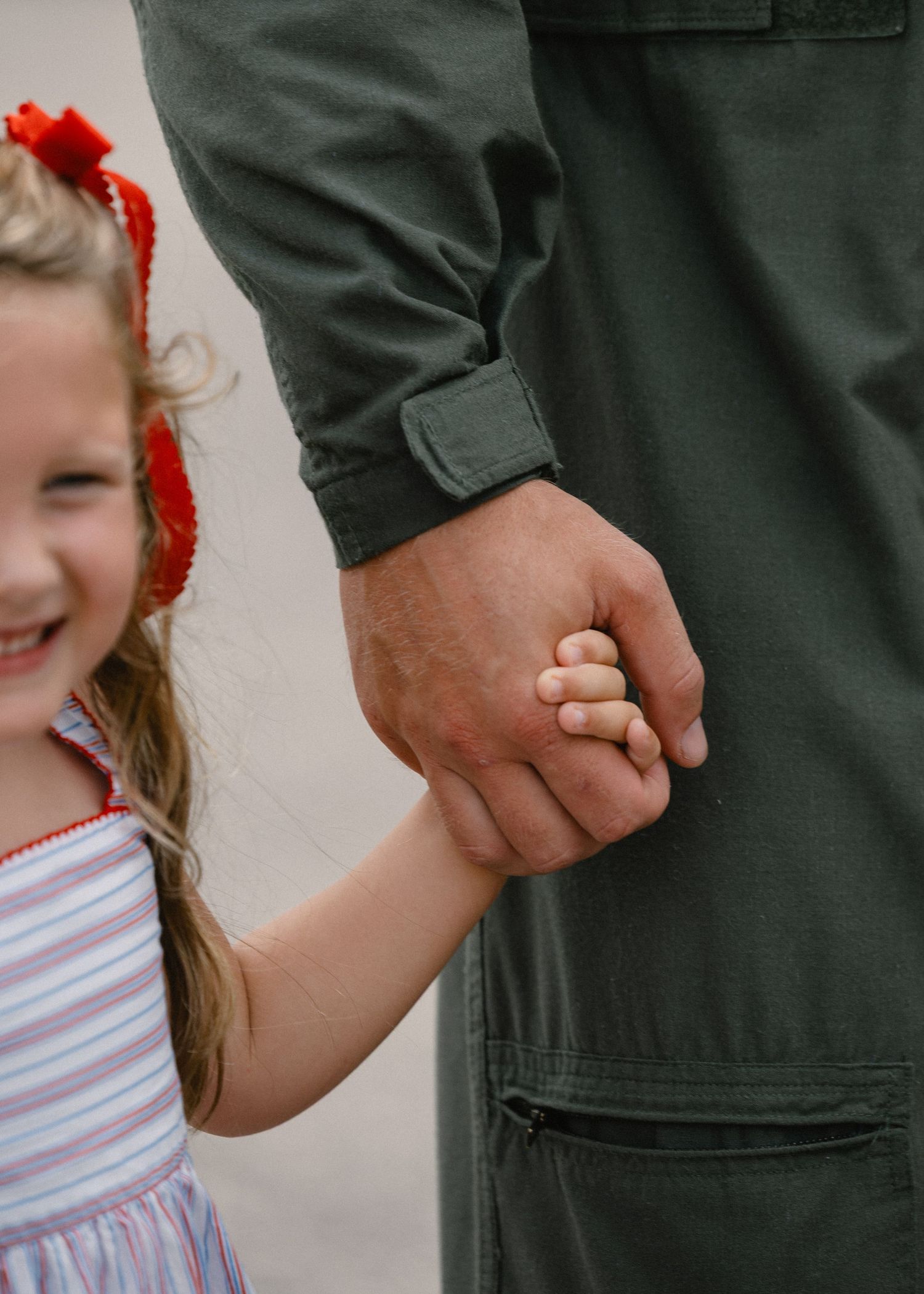 A little girl in a striped dress with red bow holds hands with a military pilot in an olive green flight suit.