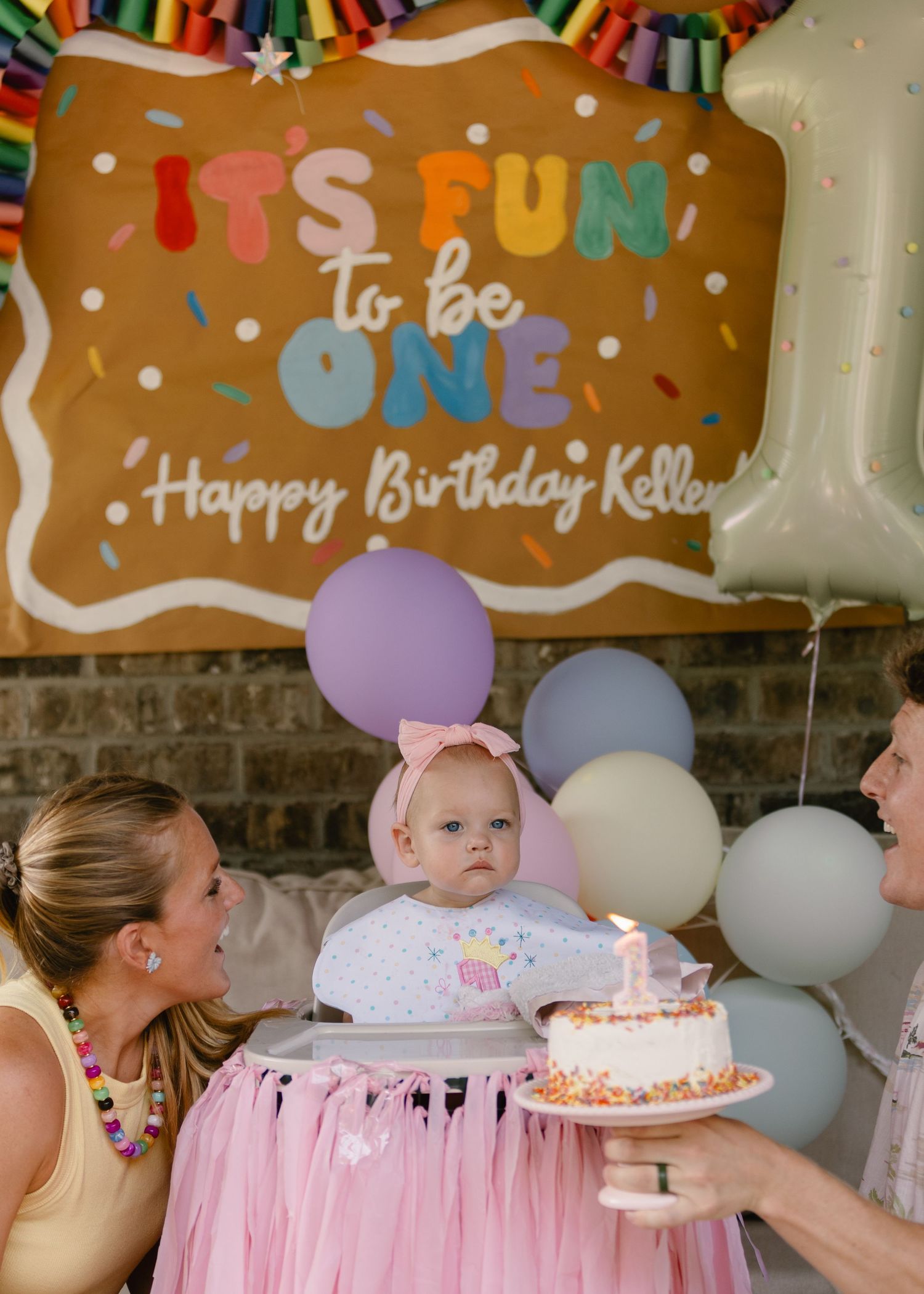 Baby girl at high chair with birthday cake surrounded by balloons and Happy Birthday Keller banner.