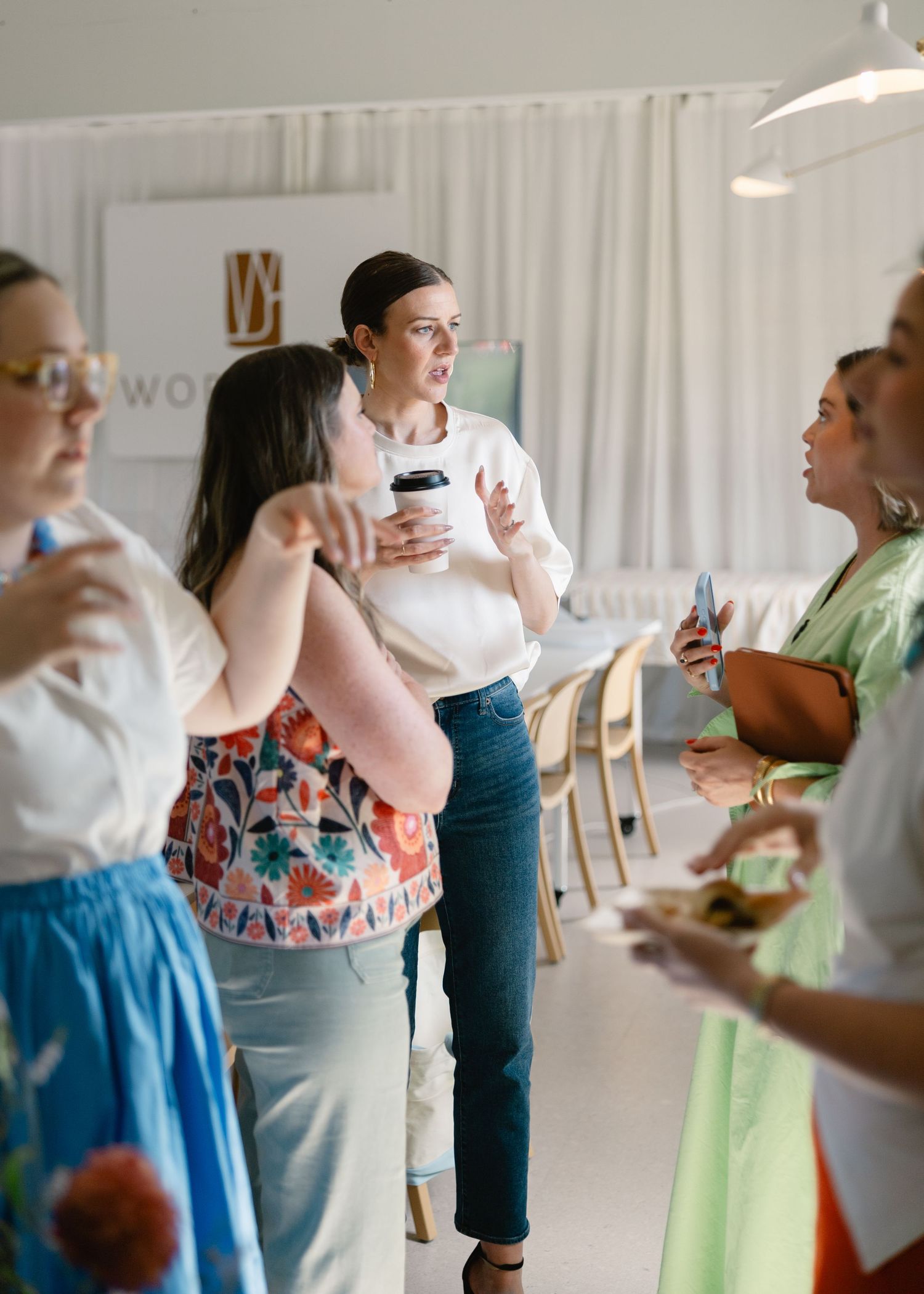 Group of women networking at a casual indoor event, holding coffee cups and plates while engaged in conversation.