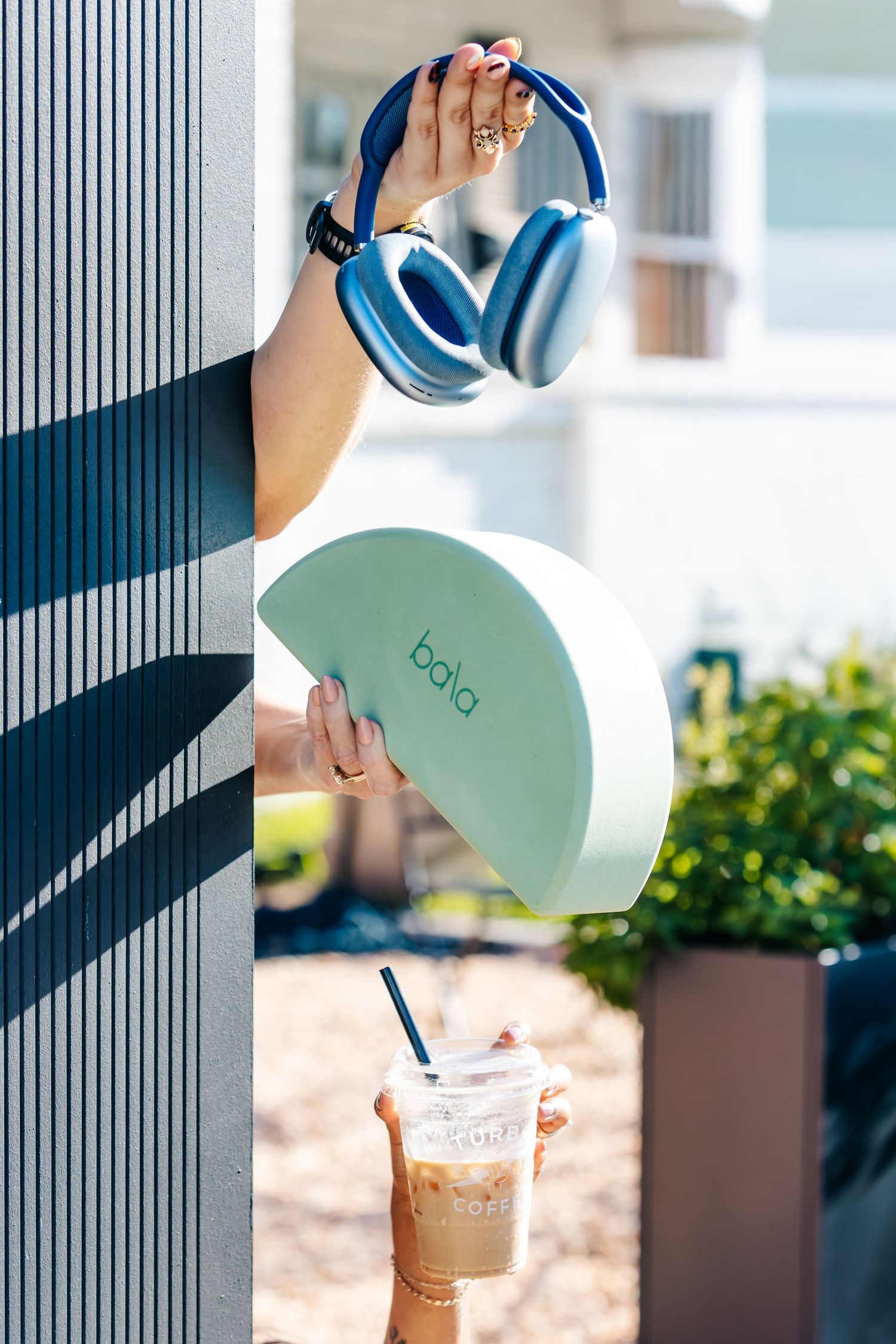 Hands holding blue headphones and a Bala yoga block with an iced coffee visible below outdoors.