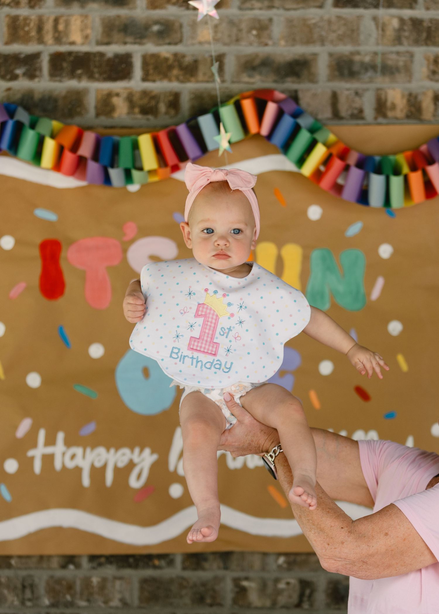 Baby in a 1st birthday bib held up in front of a Happy Birthday Keller banner on a brick wall.