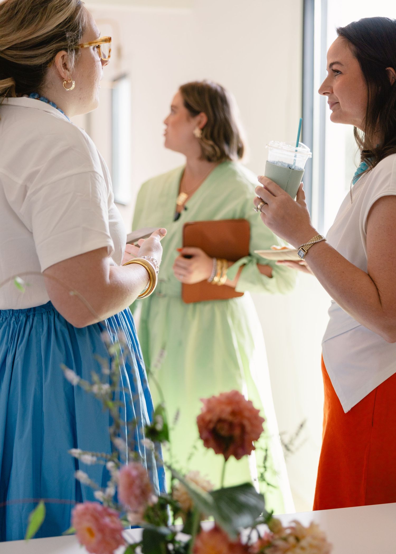 Three women socializing at an indoor event, one holding an iced drink, surrounded by colorful flowers in the foreground.