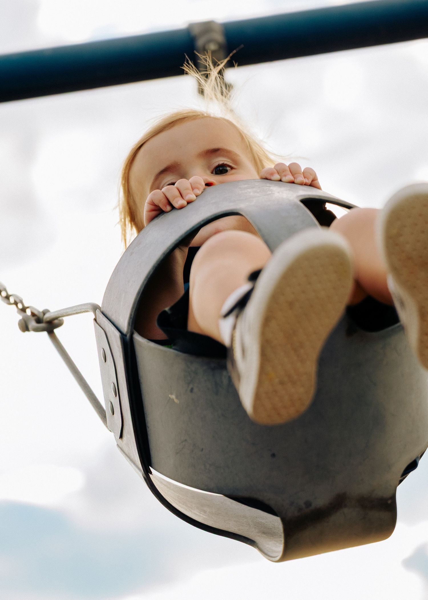 A young blonde toddler peeks over the edge of a bucket swing at a playground.