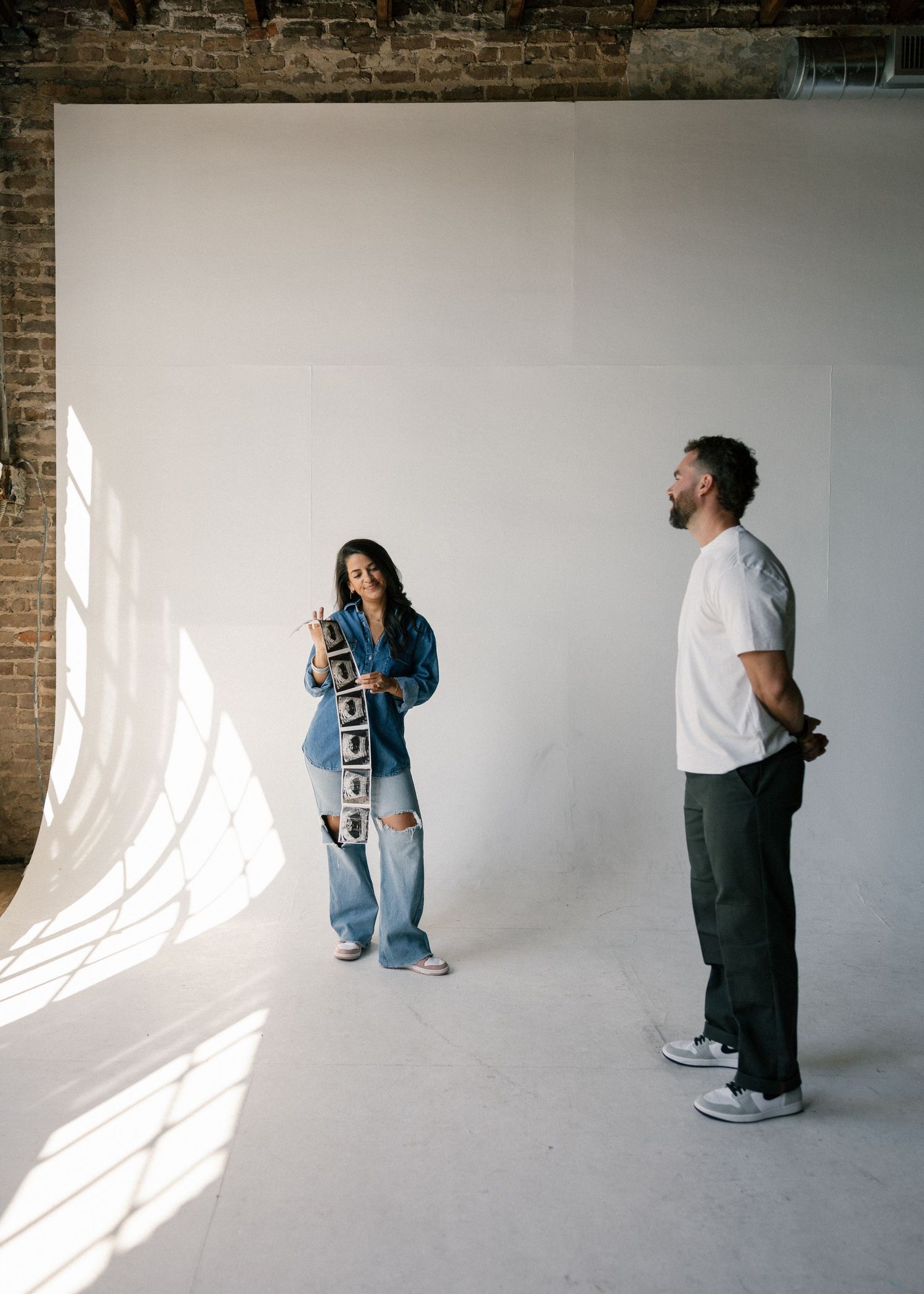 Woman shows photo strip to bearded man in bright white photography studio with brick walls.