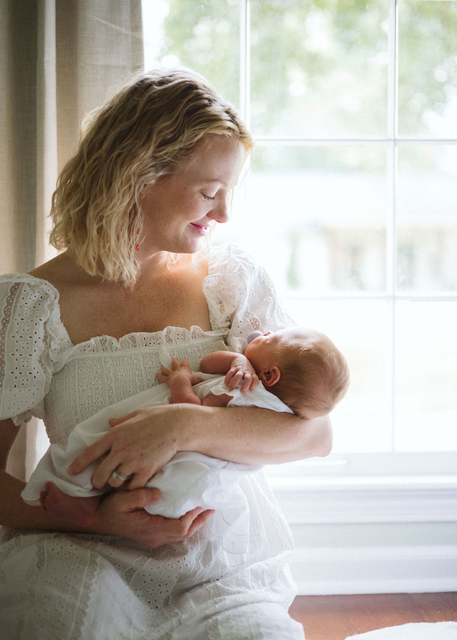 Smiling mother in a white eyelet dress cradles her sleeping newborn baby beside a sunlit window in a tender newborn portrait.