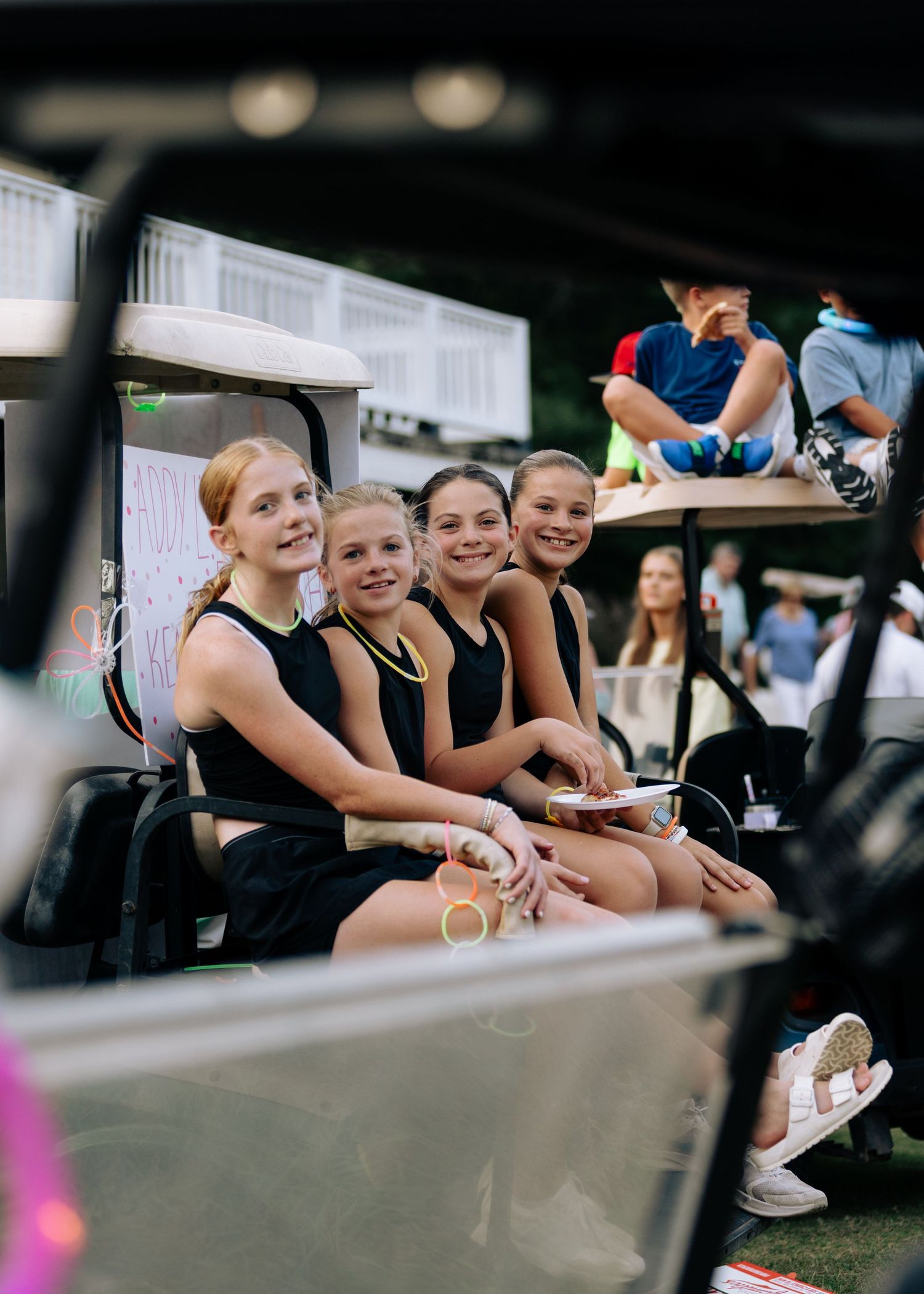 Four girls in black outfits smile while seated together on a golf cart at an outdoor event.