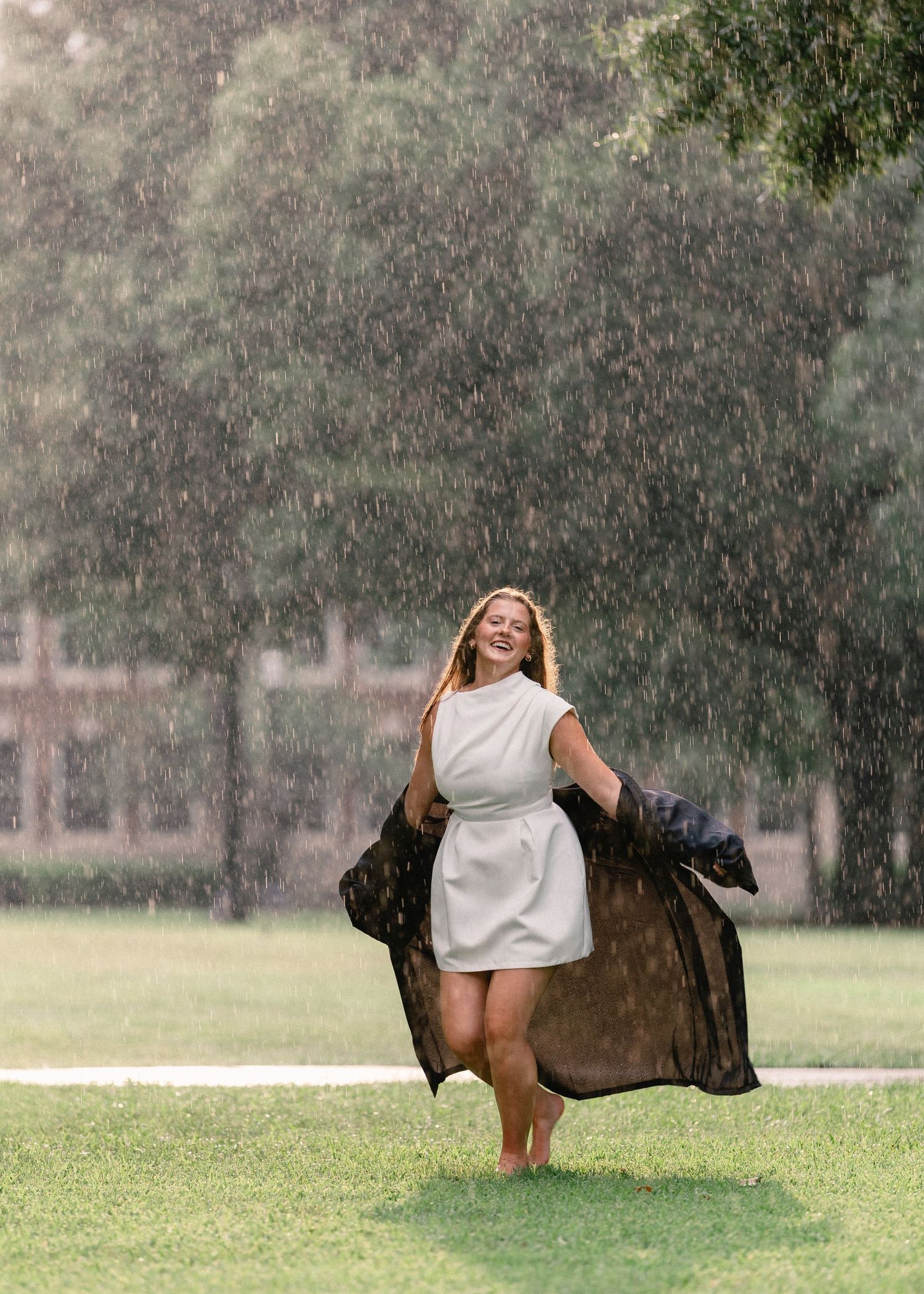 Joyful graduate laughs in rain wearing white dress holding black gown on green campus.