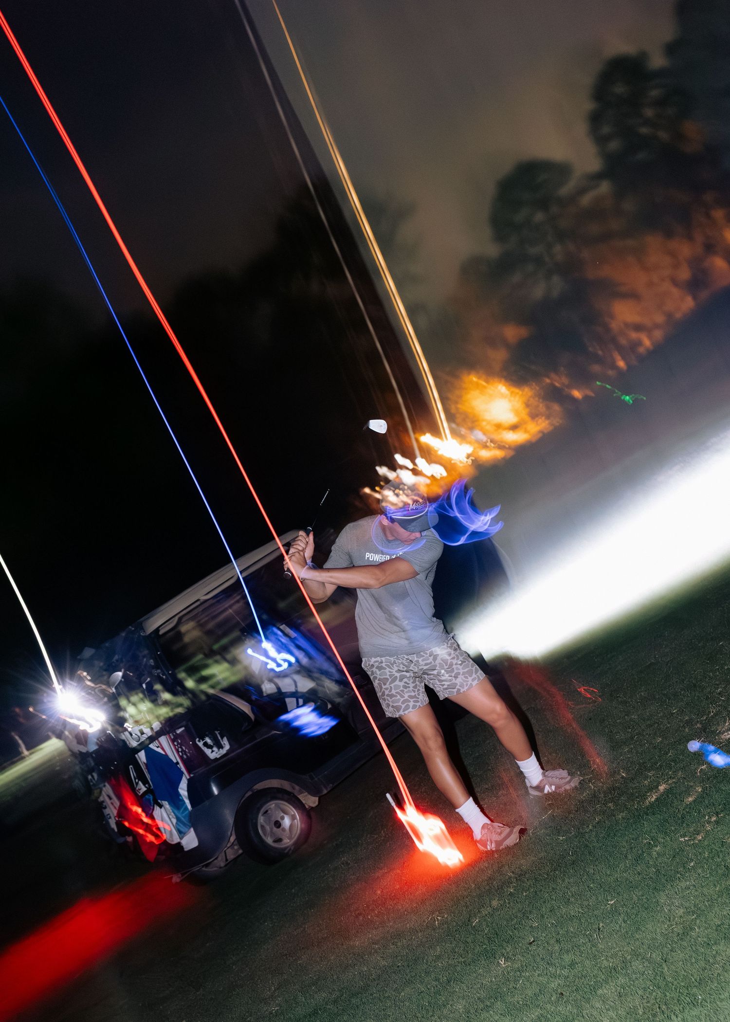 Golfer swings with vibrant red and blue light trails streaking across a dark night sky.