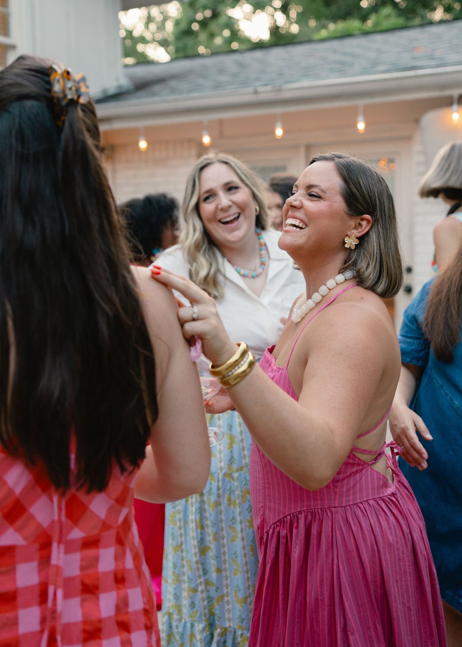 Two women laughing and dancing together at a festive outdoor evening party with string lights behind them.