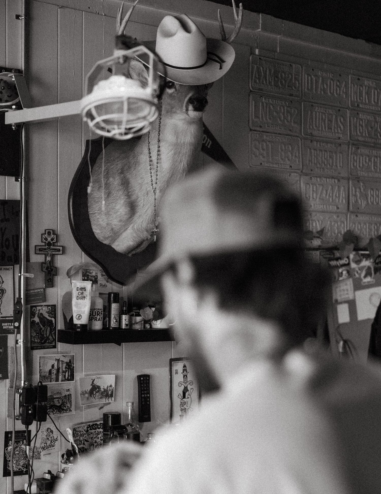 Black and white photo of a man in a cowboy hat near a mounted deer head in a rustic shop.