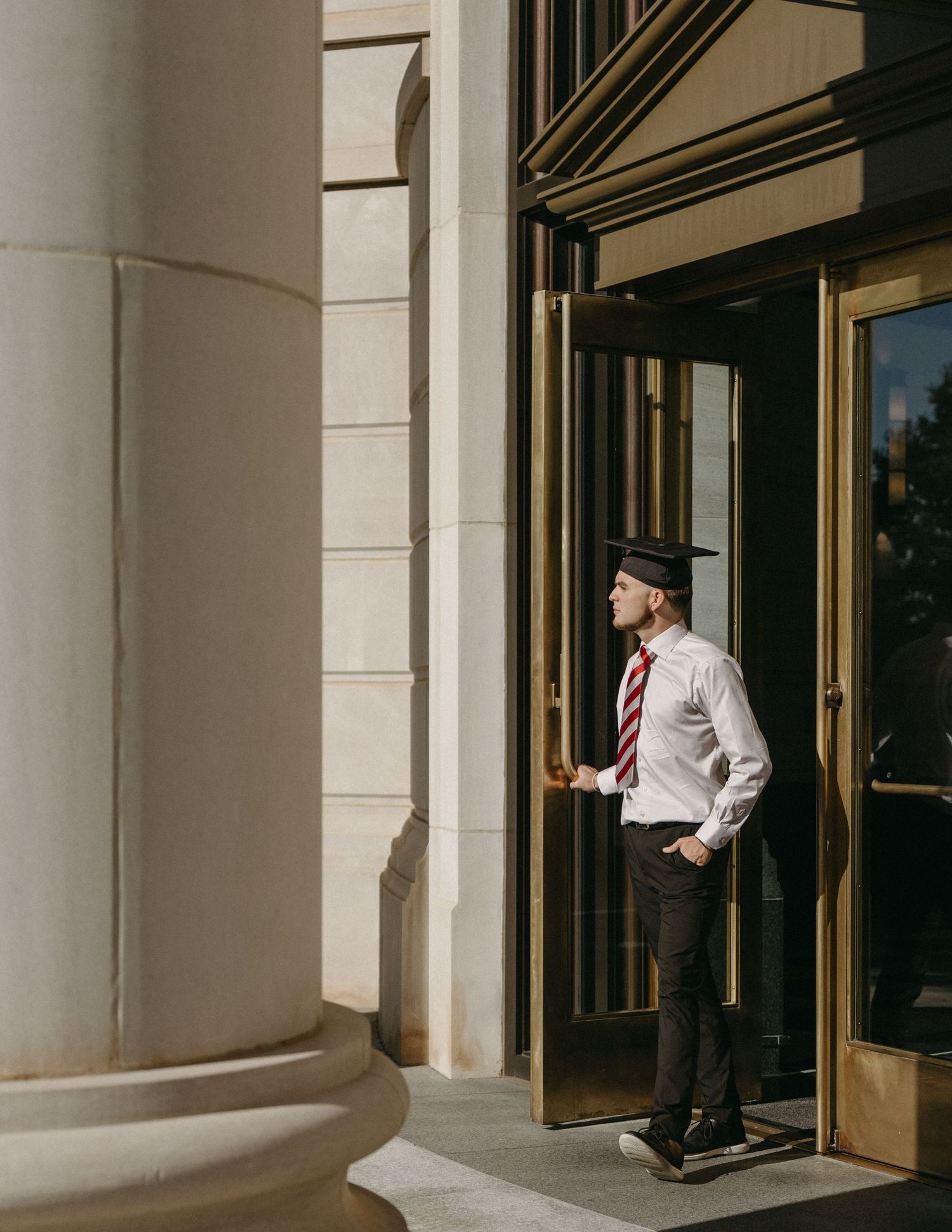 Graduate in cap and tie opening golden revolving door of classical building with large columns.