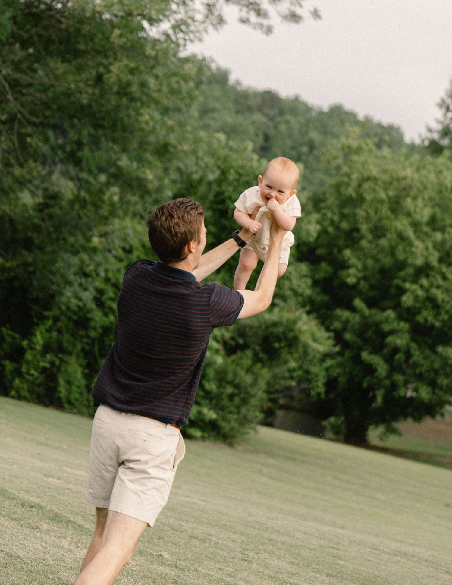 Father lifting happy baby high in the air outdoors on a lush green lawn.
