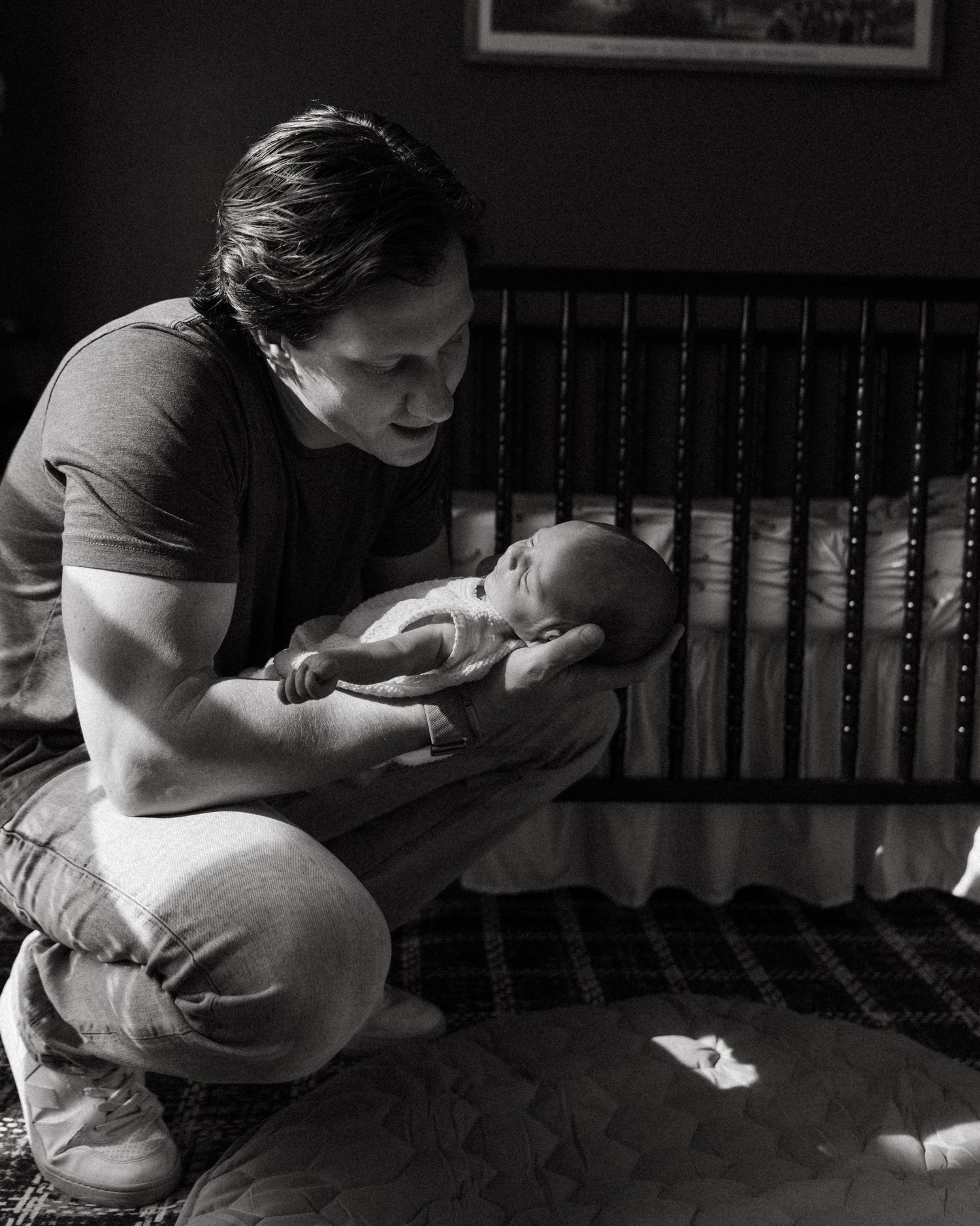 Black and white photo of father kneeling beside crib holding newborn baby.