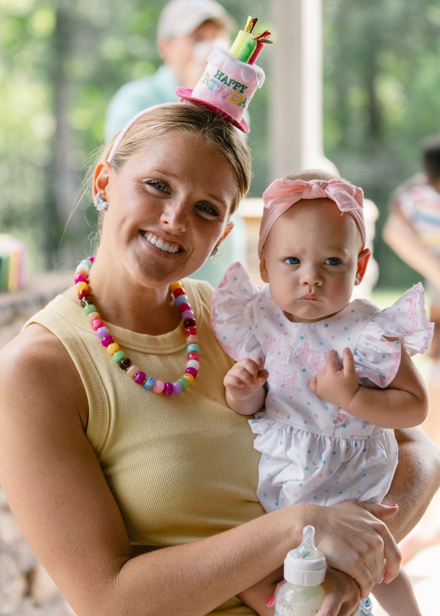 Smiling woman wearing a birthday cake hat and colorful bead necklace holds a baby girl in a pink bow headband.