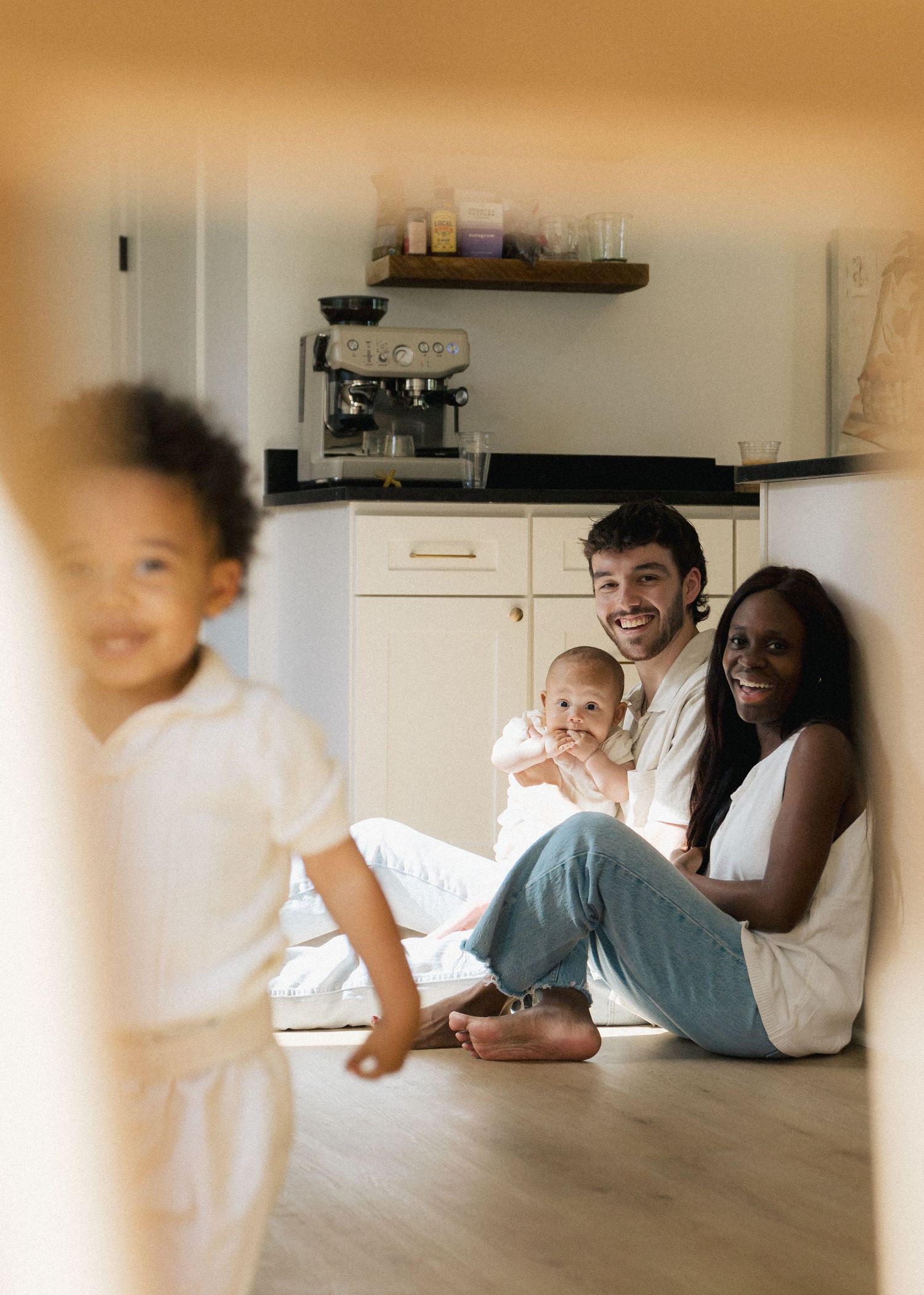 An interracial family sits on the kitchen floor smiling with their baby and toddler nearby.