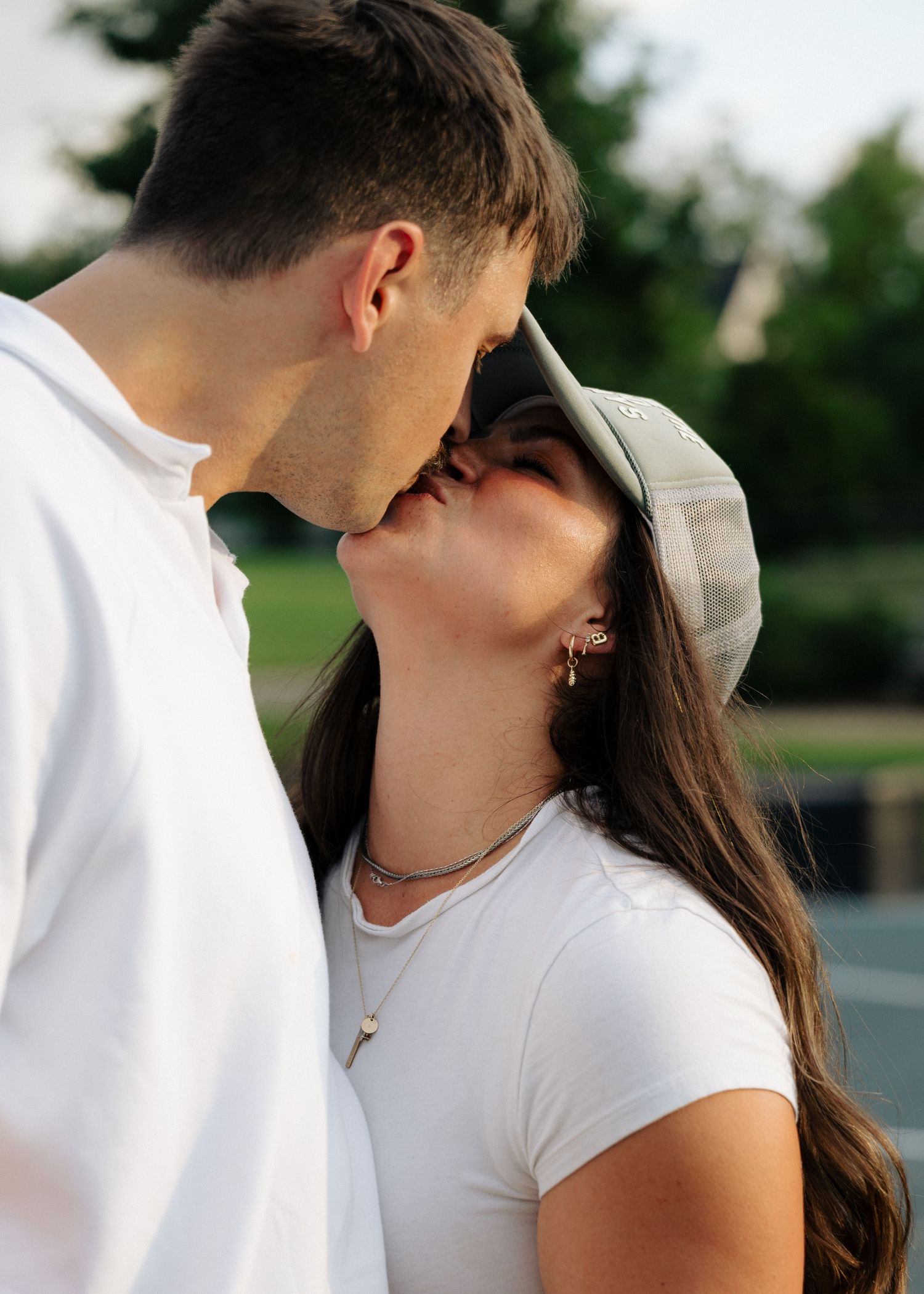 A couple shares a romantic kiss outdoors in warm golden sunlight, the woman wearing a gray cap.