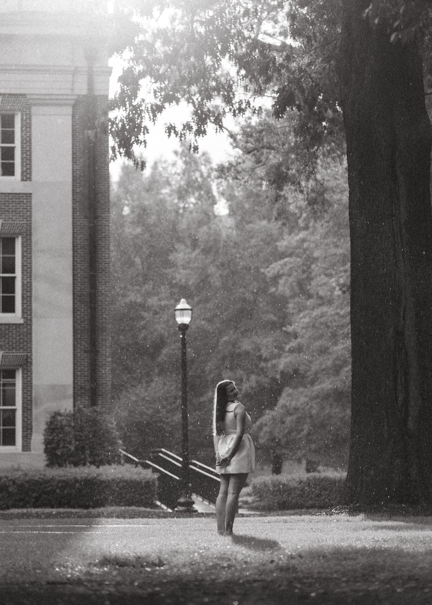 Graduate stands alone in rain near lamp post and brick building on college campus.