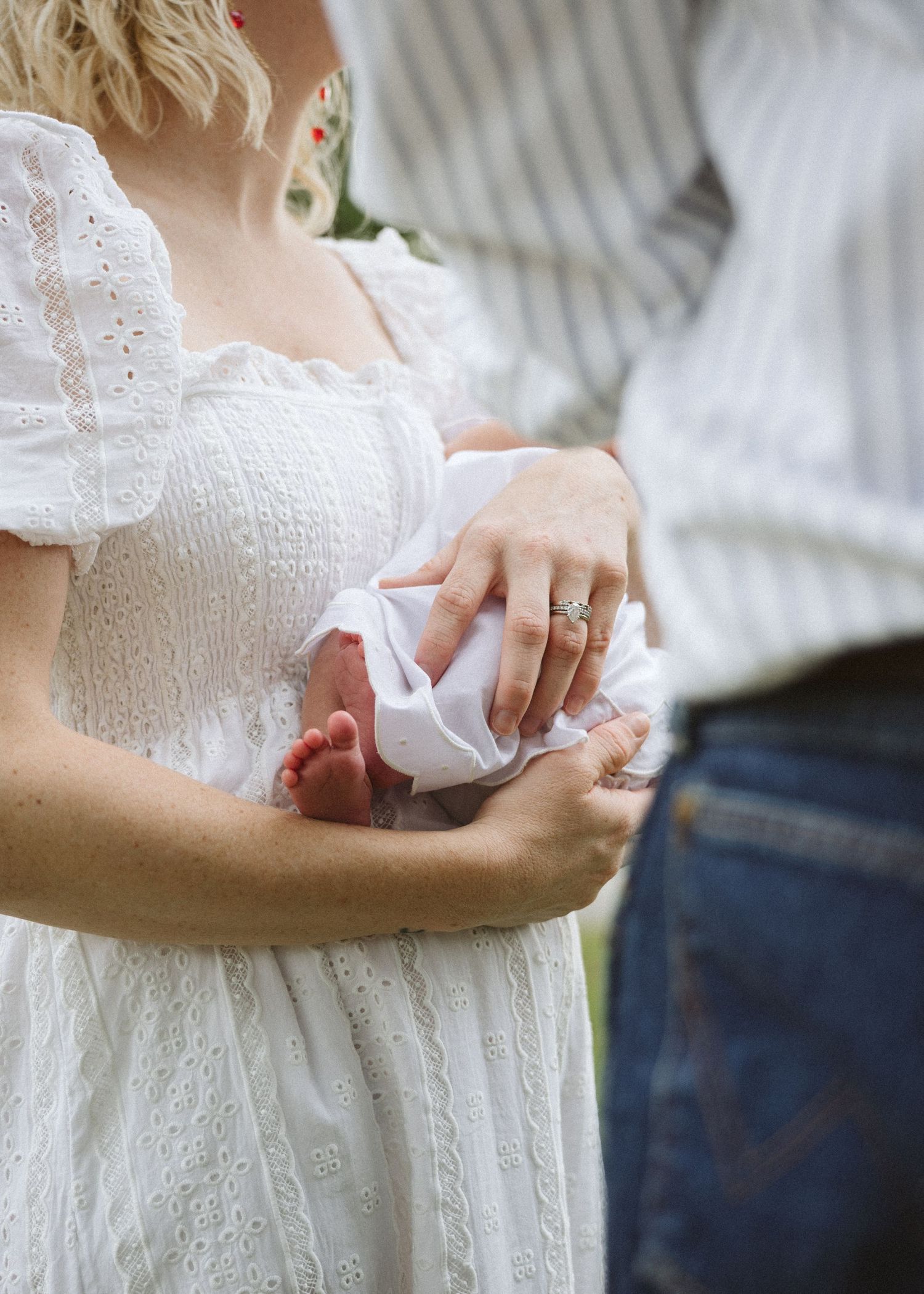 Woman in a white eyelet dress cradling a swaddled newborn with tiny bare feet visible while a man in jeans stands beside her.