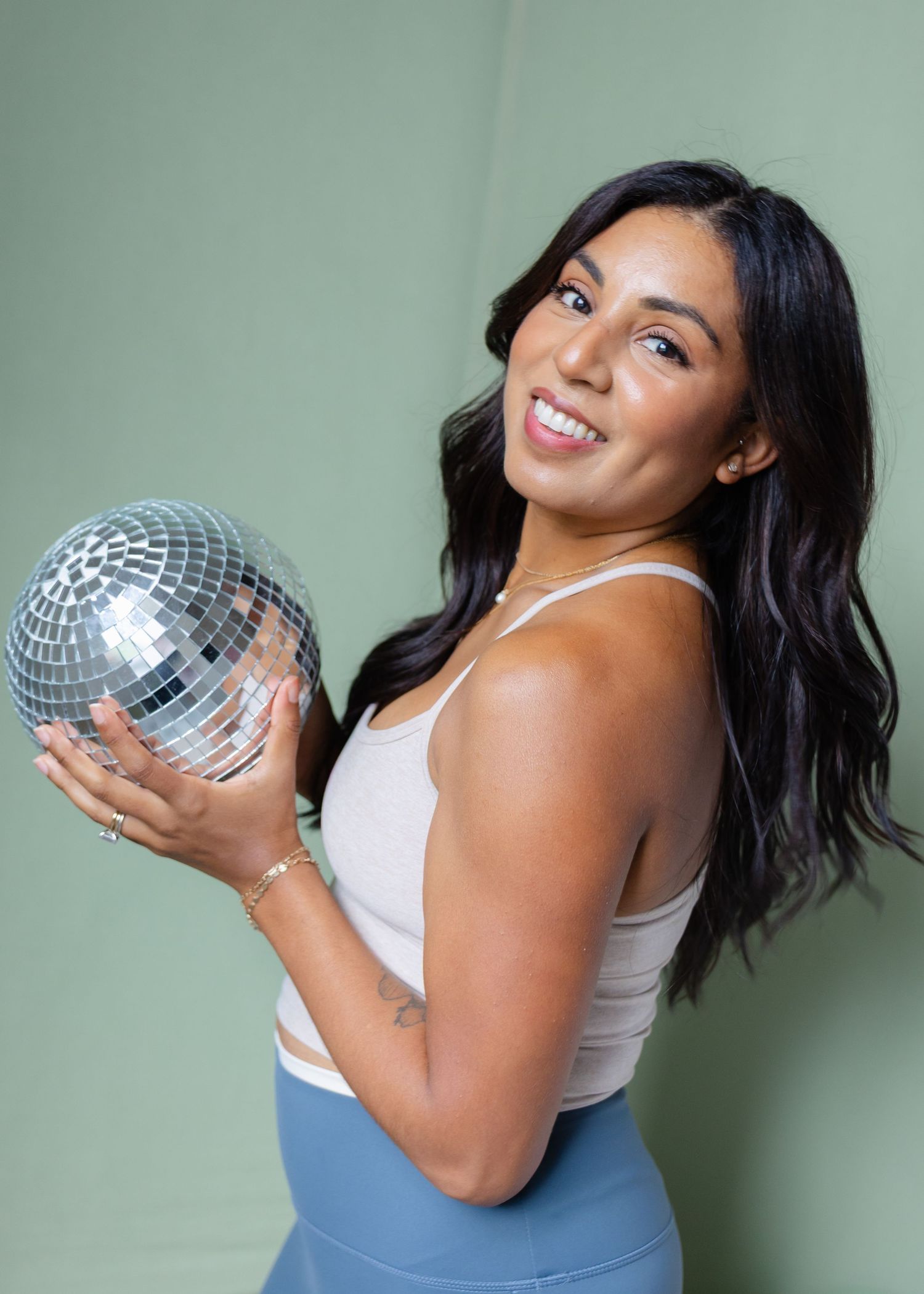 Smiling woman in beige tank top holding silver disco ball against green background.