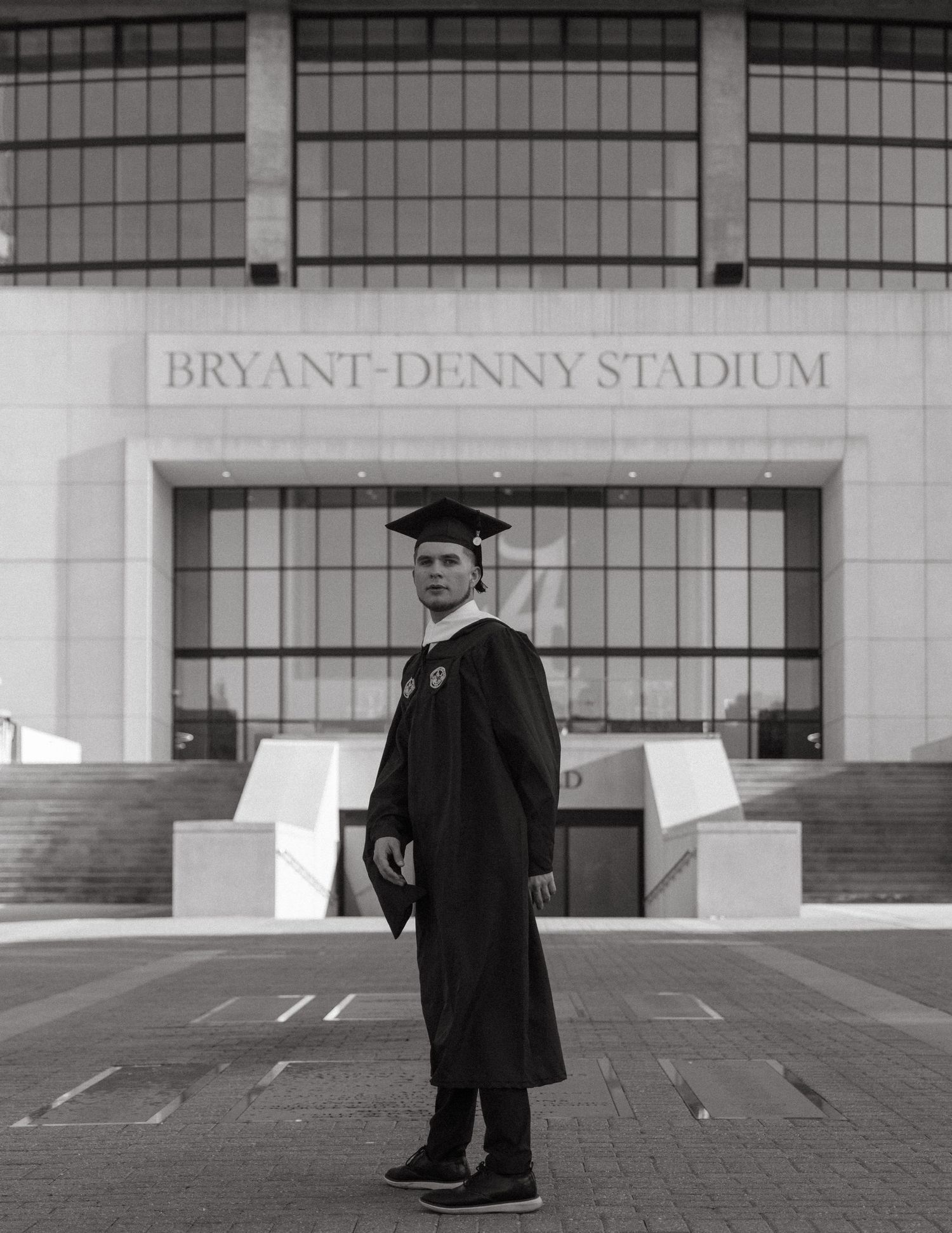 Graduate in cap and gown posing in front of Bryant-Denny Stadium in black and white photo.