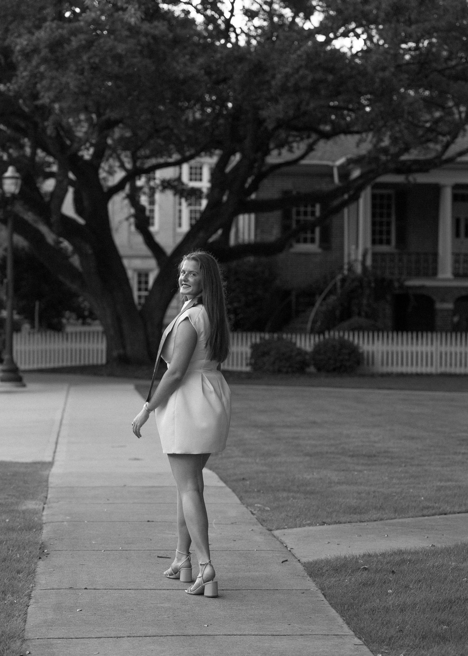 Black and white portrait of young woman in white dress smiling on sidewalk near historic building.