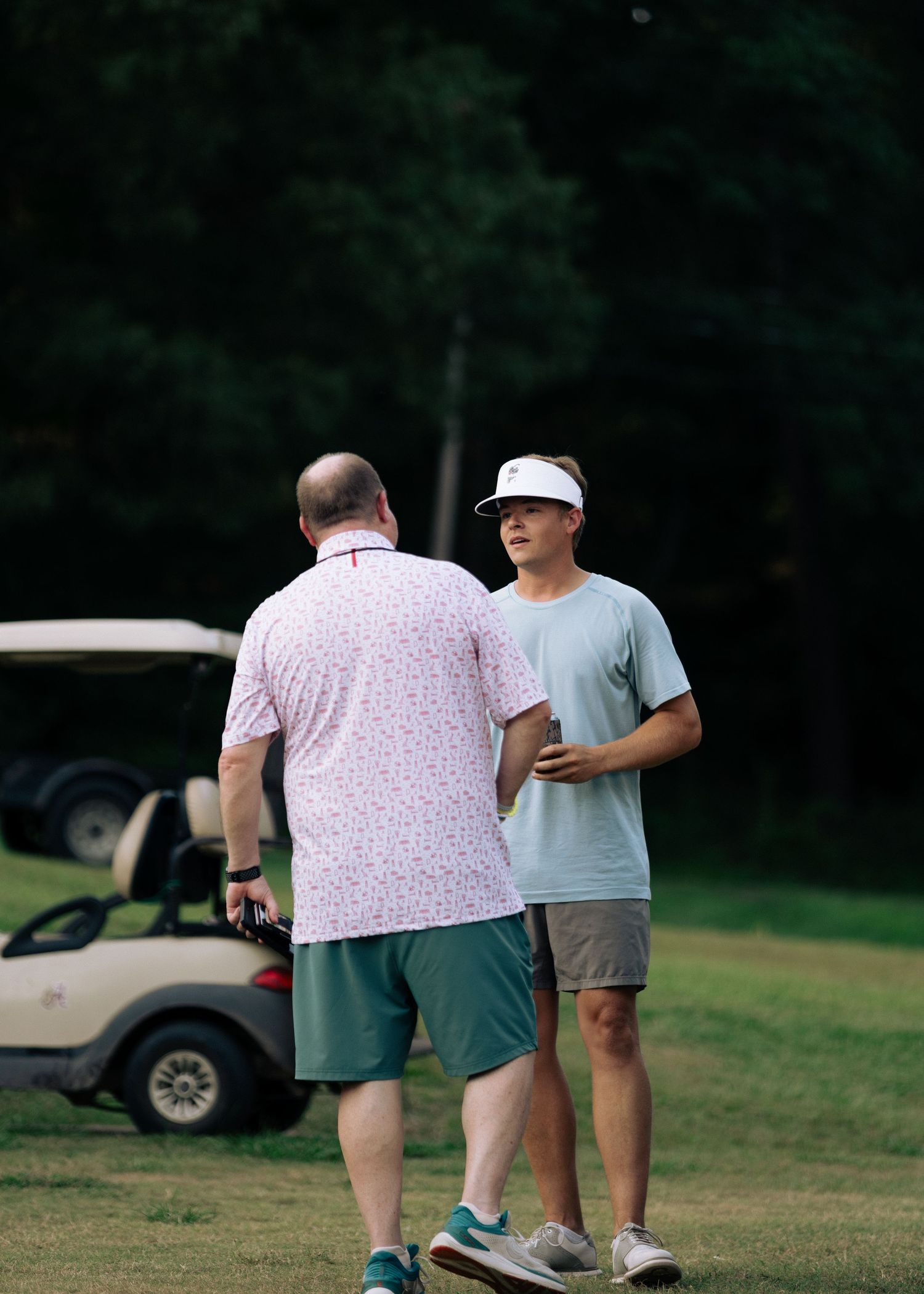 Two men conversing on a golf course with golf carts parked in background.