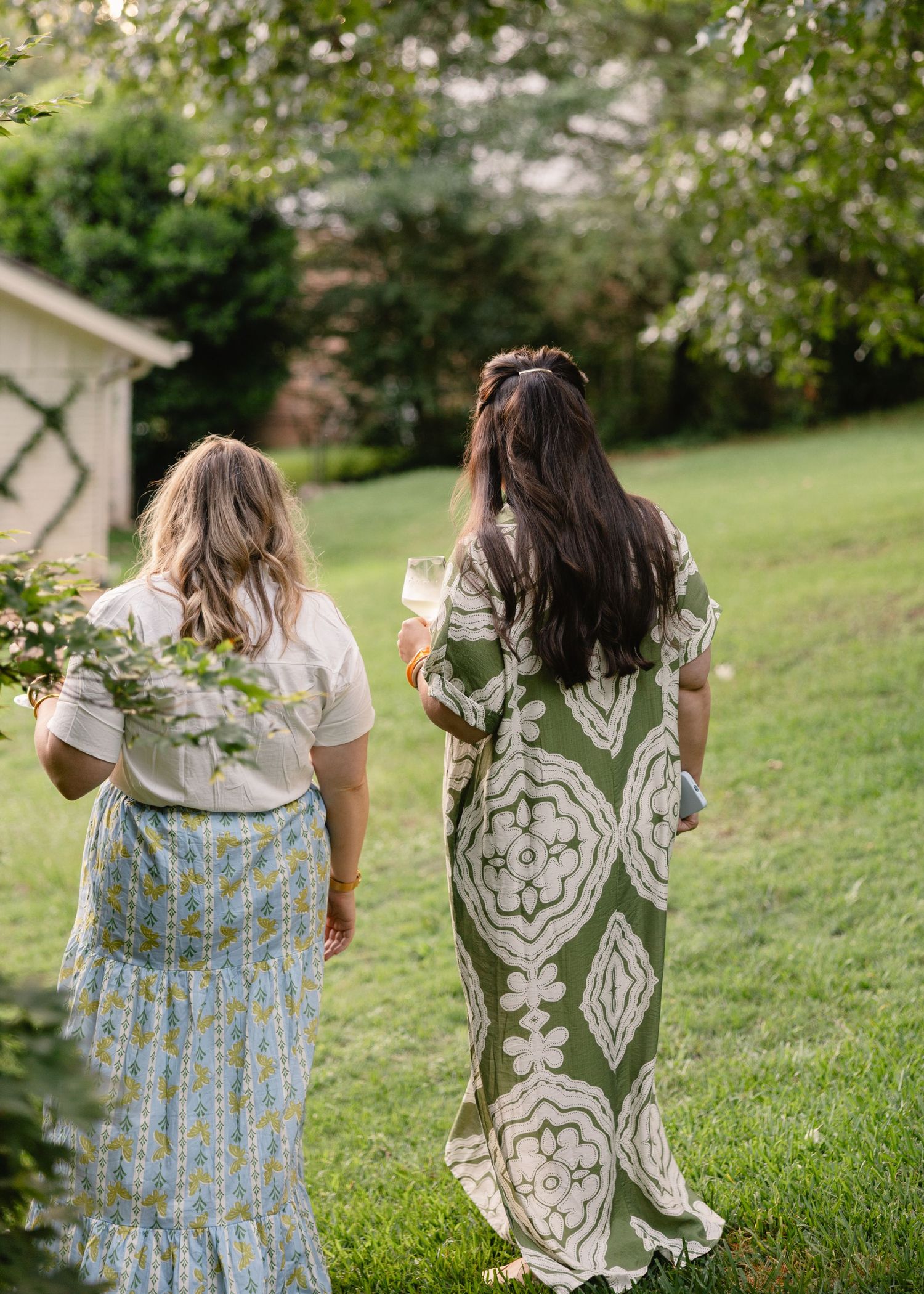 Two women walking through lush green garden, one in floral dress, one in olive maxi.
