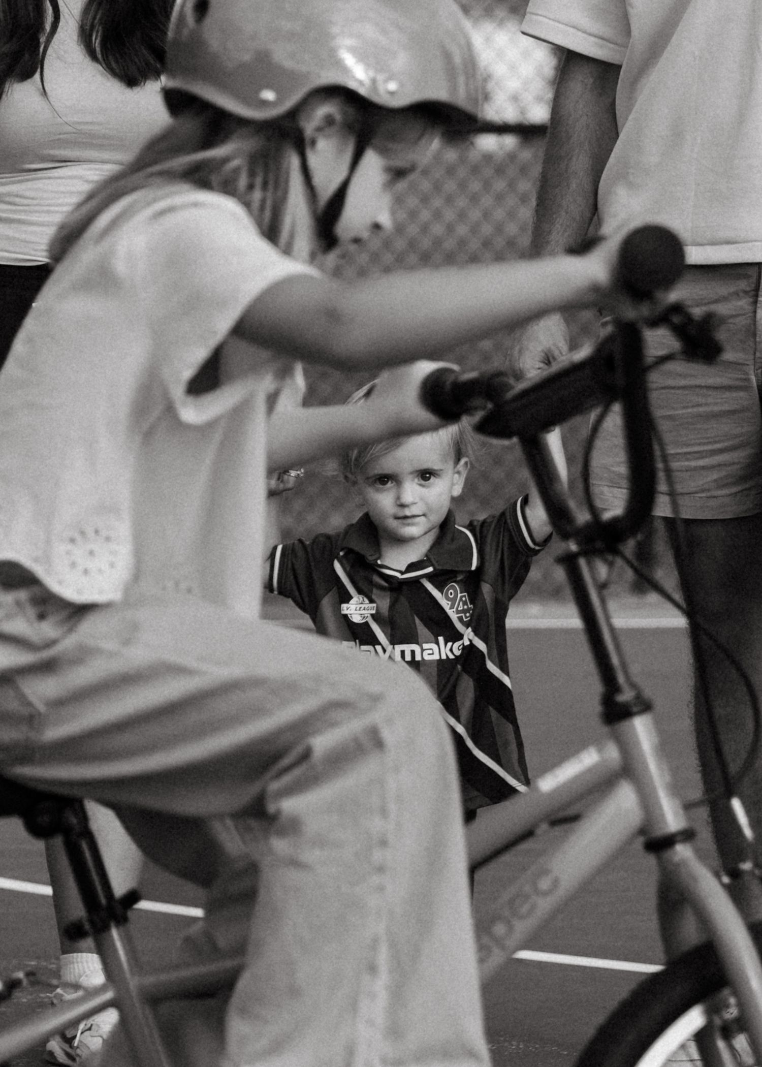 A young boy in a soccer jersey watches a girl on a bike at an indoor court in black and white.