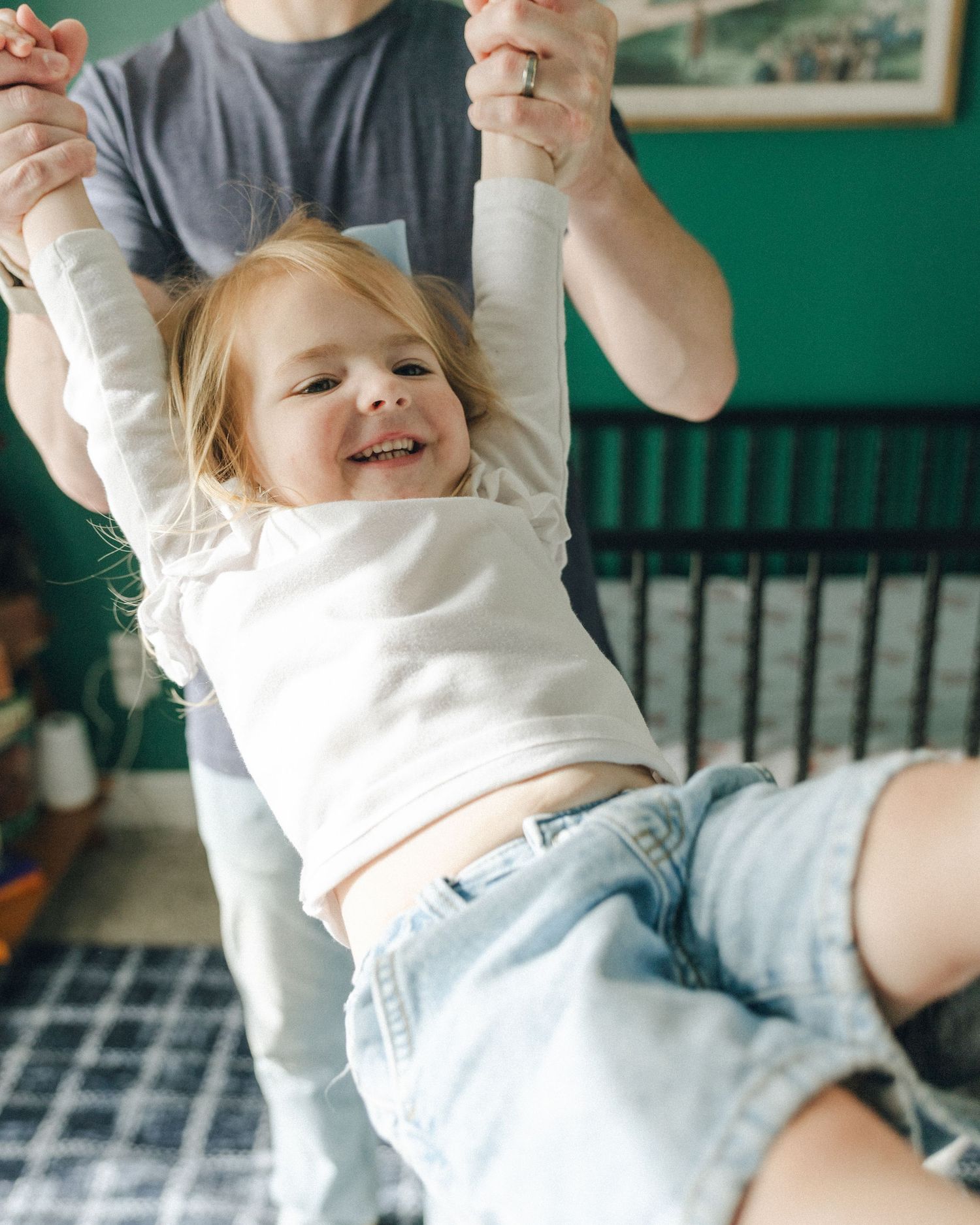 Laughing toddler girl being lifted by parent in colorful nursery room with green walls and crib.