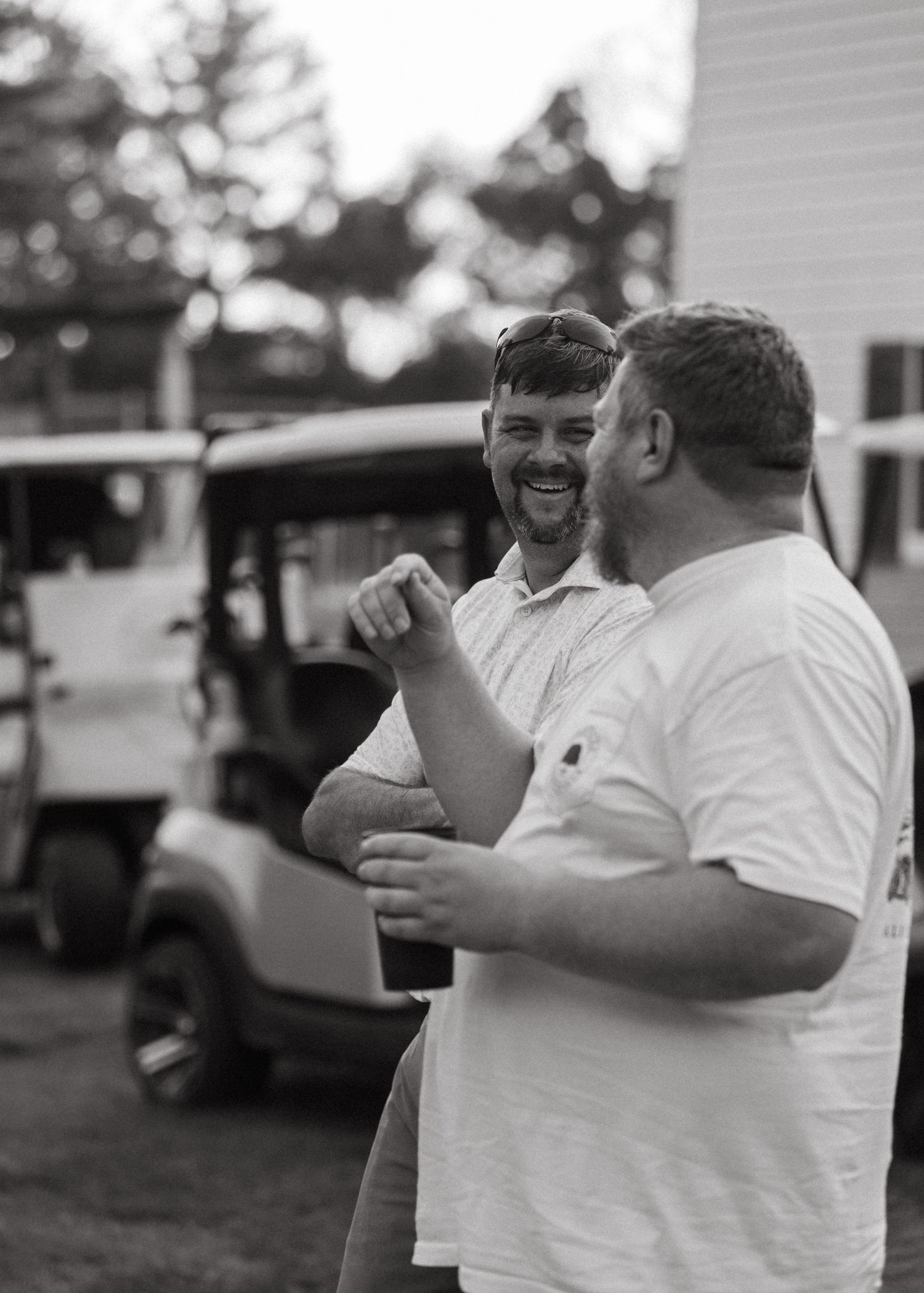 Two men laughing and talking together near white golf carts in black and white.