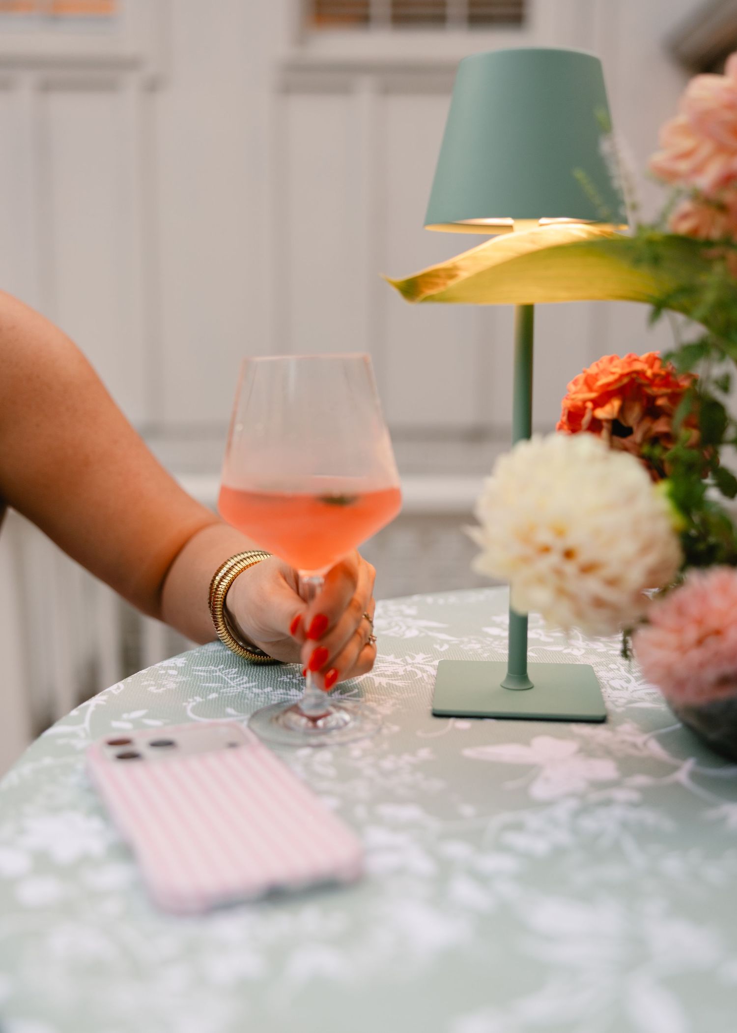 Hand with gold bracelets holding rose wine glass near floral centerpiece and teal lamp.