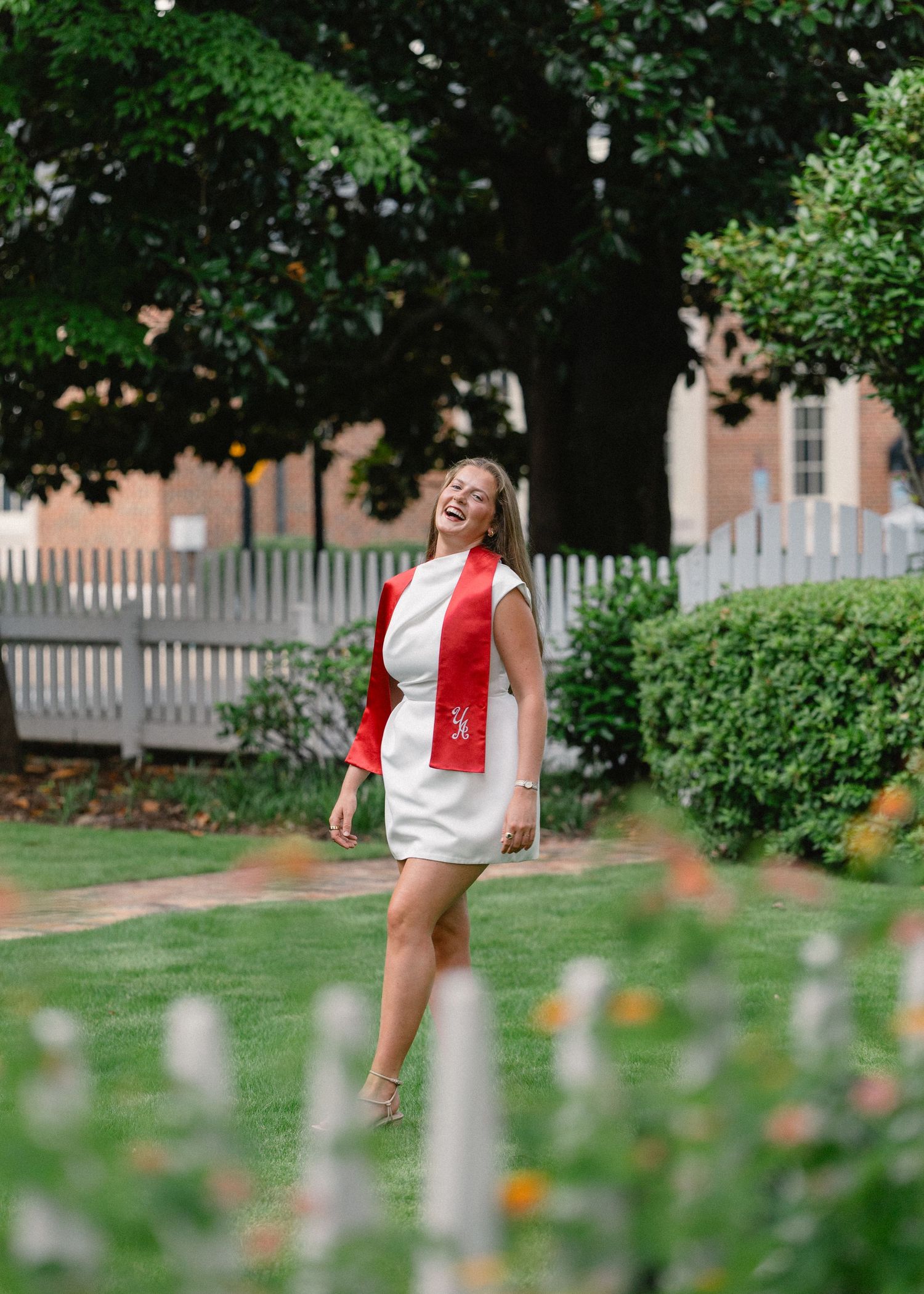 Laughing graduate in white dress and red graduation stole walking through garden near brick building.