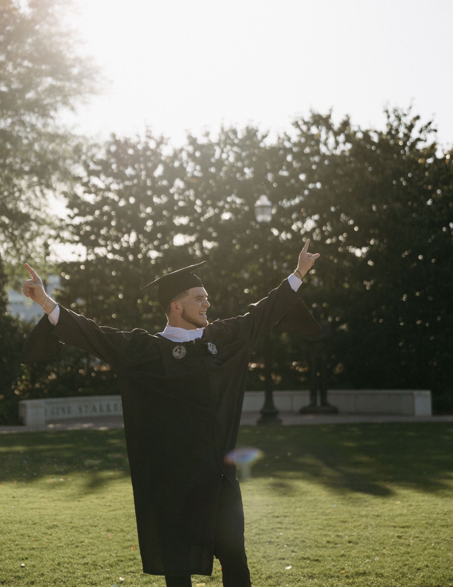 Joyful graduate in cap and gown celebrating with arms raised wide on sunny campus lawn.