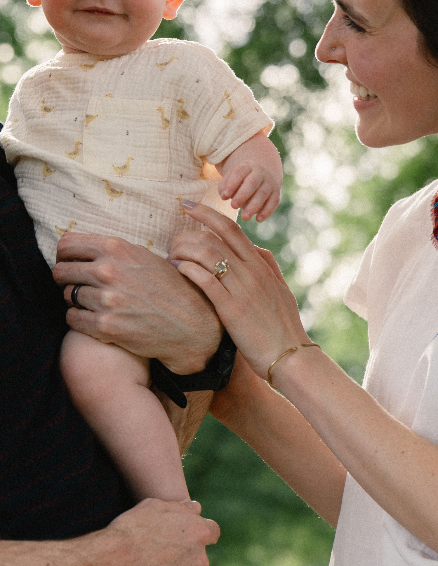 Close-up of smiling mother reaching for baby in duck-print onesie held by father.