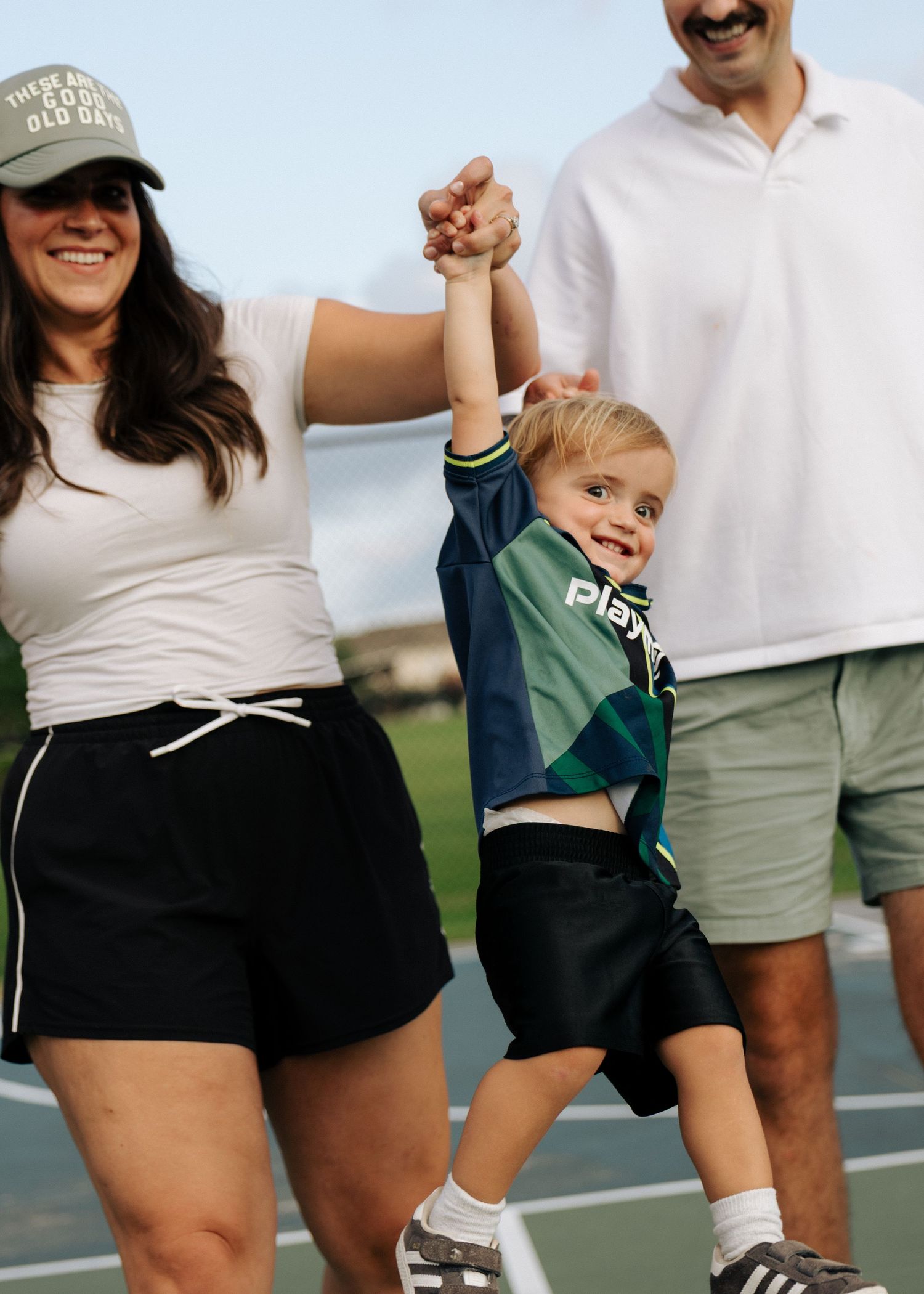 Two parents swing a smiling toddler in a colorblock jersey between them on a tennis court.