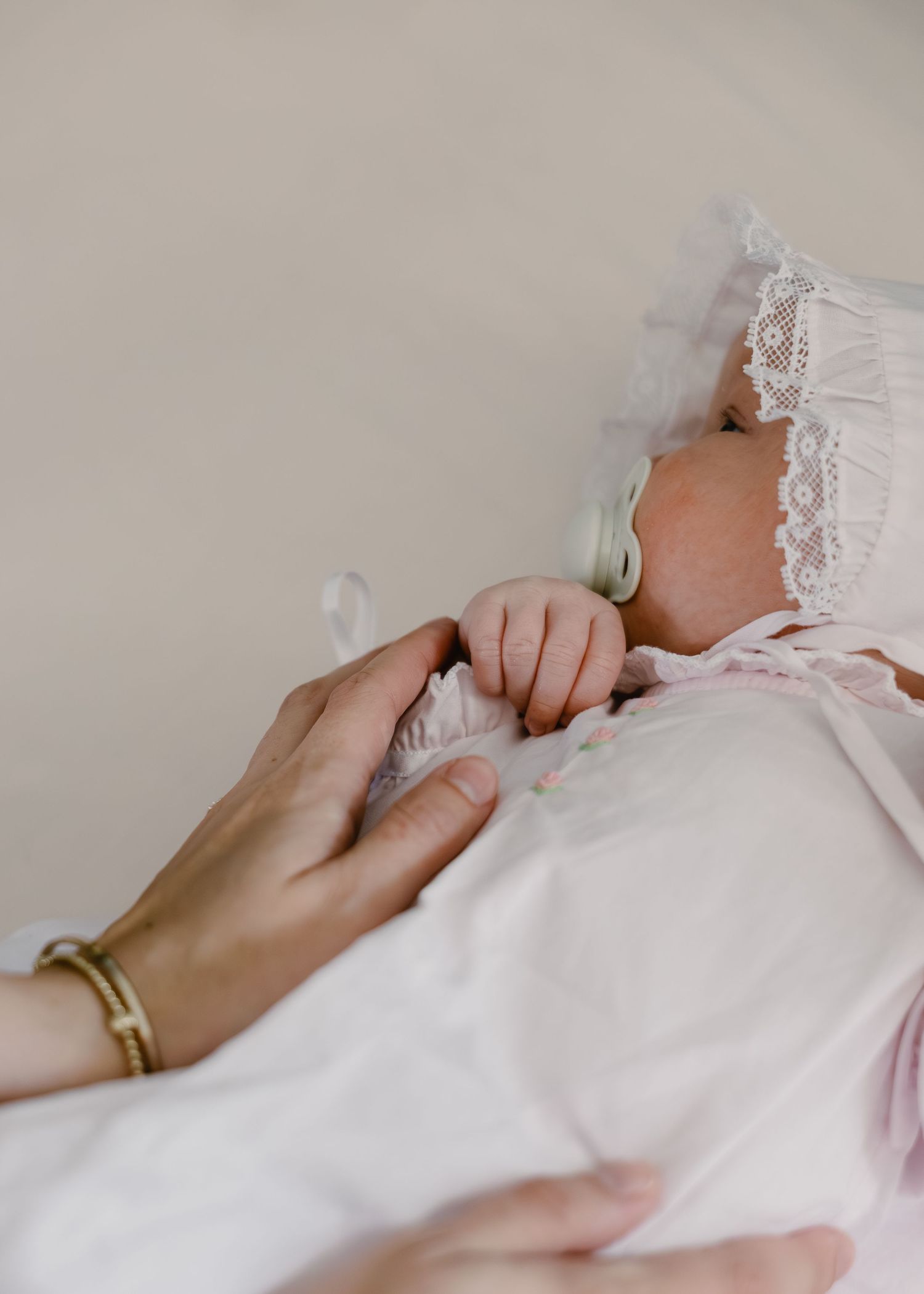 Mother's hand holding newborn baby girl in white lace bonnet and pink outfit.