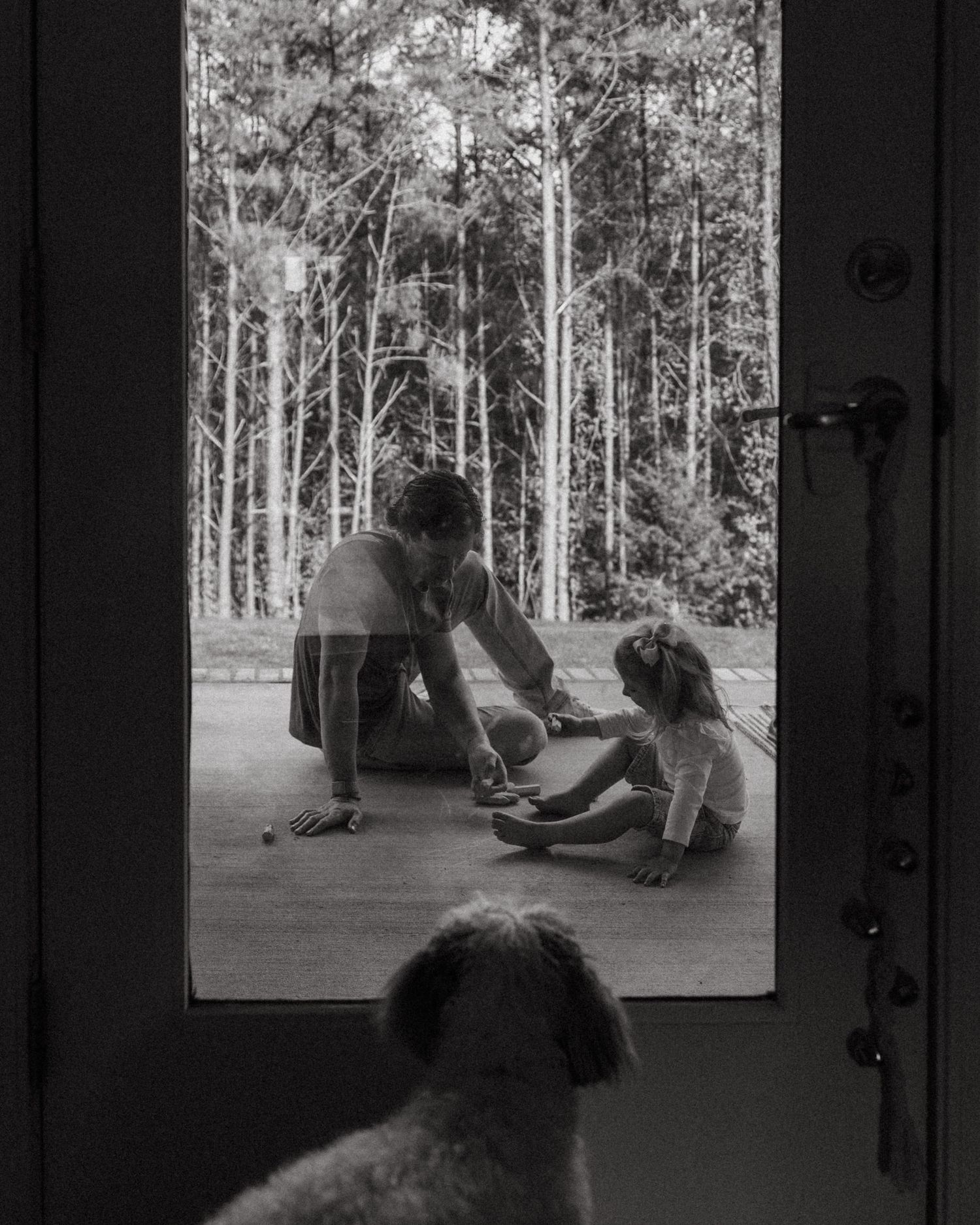 Black and white photo of parent playing with young child on porch, viewed through door with dog watching.