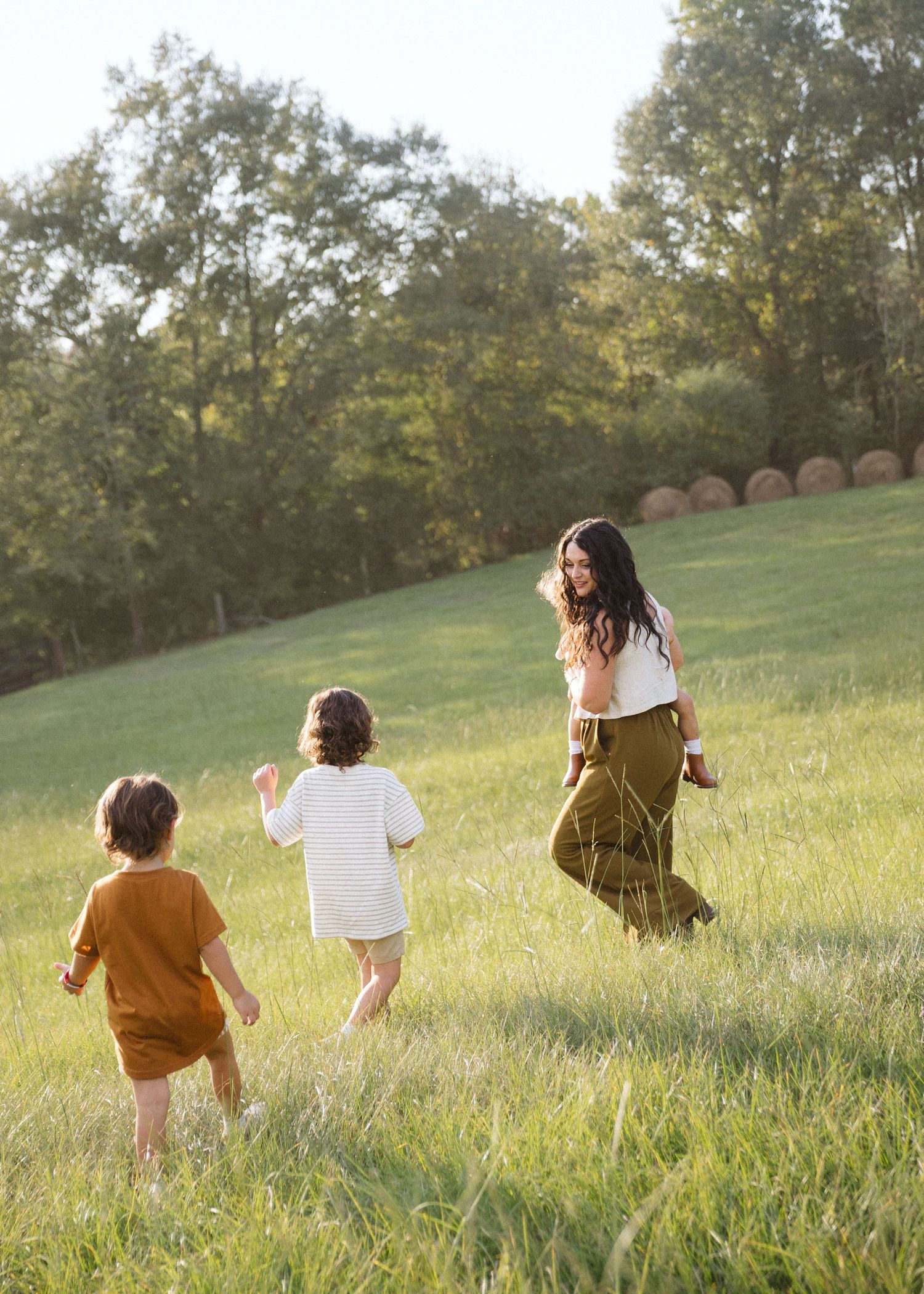 A woman and two young children in earthy tones run through a sunlit grassy hillside with hay bales and trees in the background.