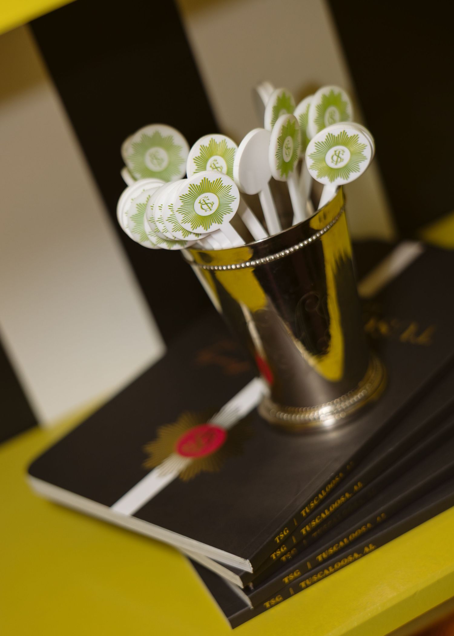 Silver julep cup holding green and white monogrammed drink stirrers, resting on a stack of black books atop a yellow table.
