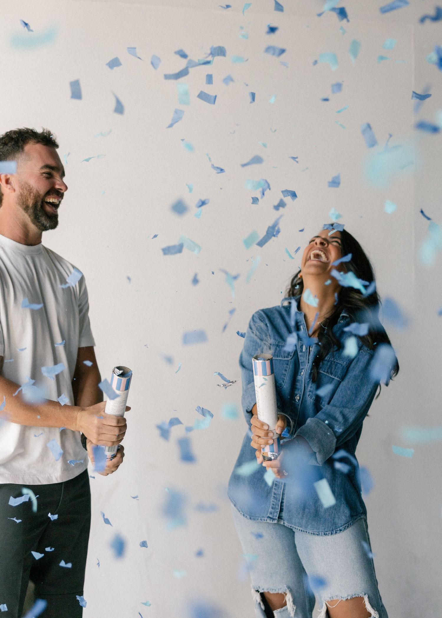 Couple celebrating with blue confetti cannons during a joyful baby boy gender reveal photoshoot.