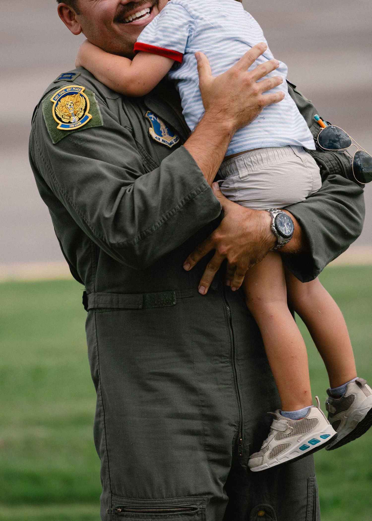 Smiling military pilot in green flight suit with unit patch joyfully holds a young toddler during homecoming reunion.