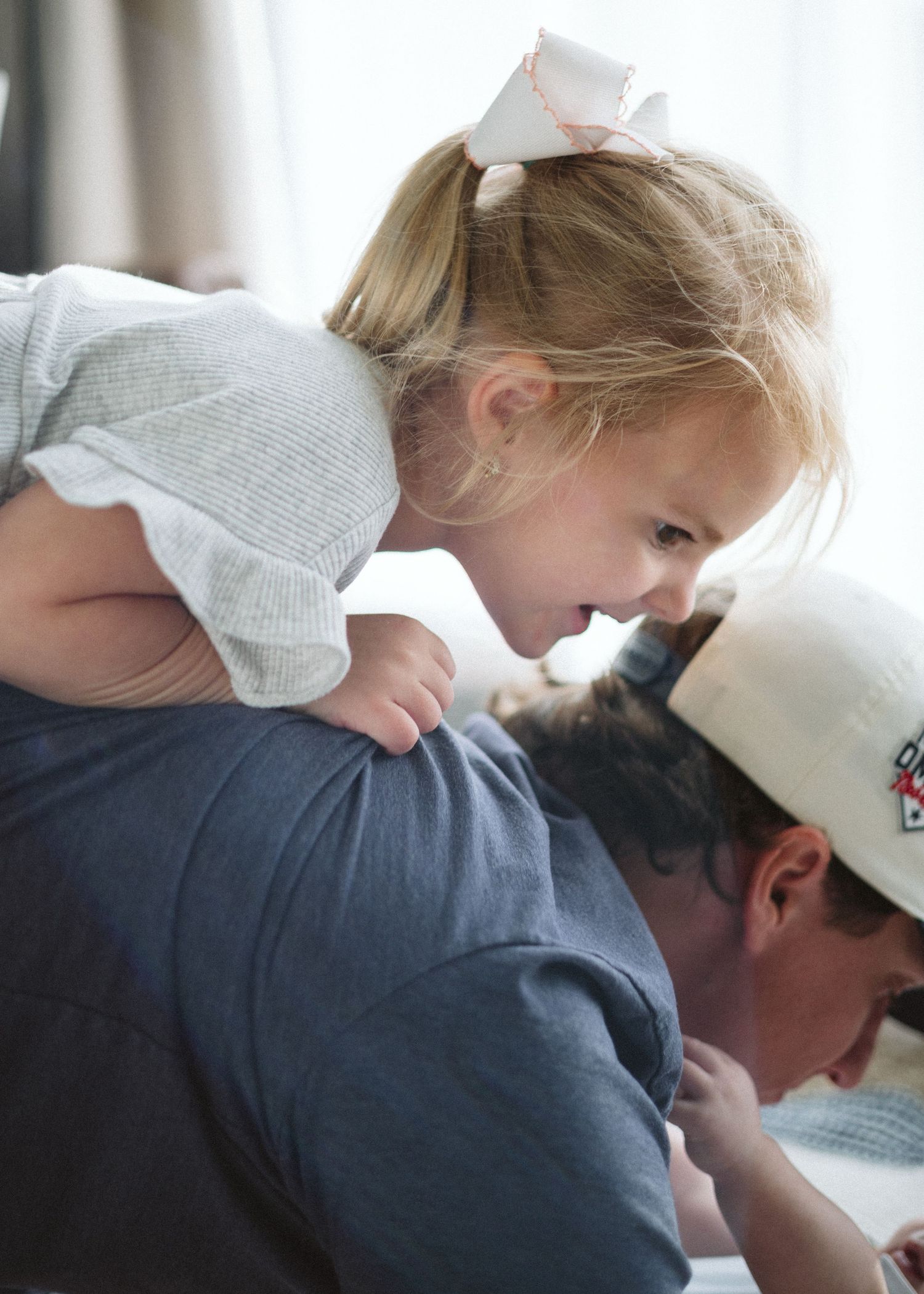 Young toddler girl with a white bow rides on an adult's shoulders during playtime.