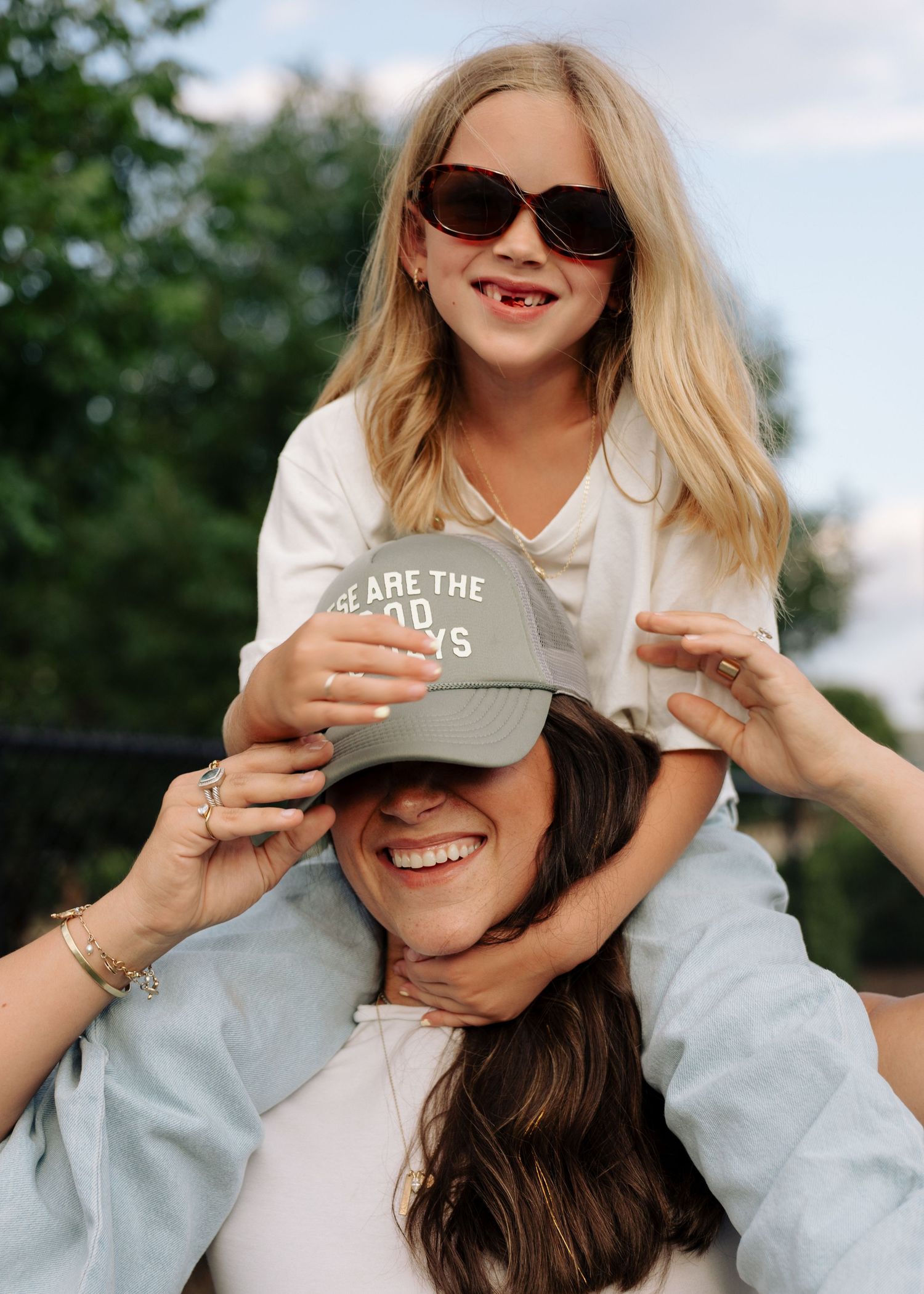 A young girl sits on a womans shoulders outdoors, both smiling on a sunny day.