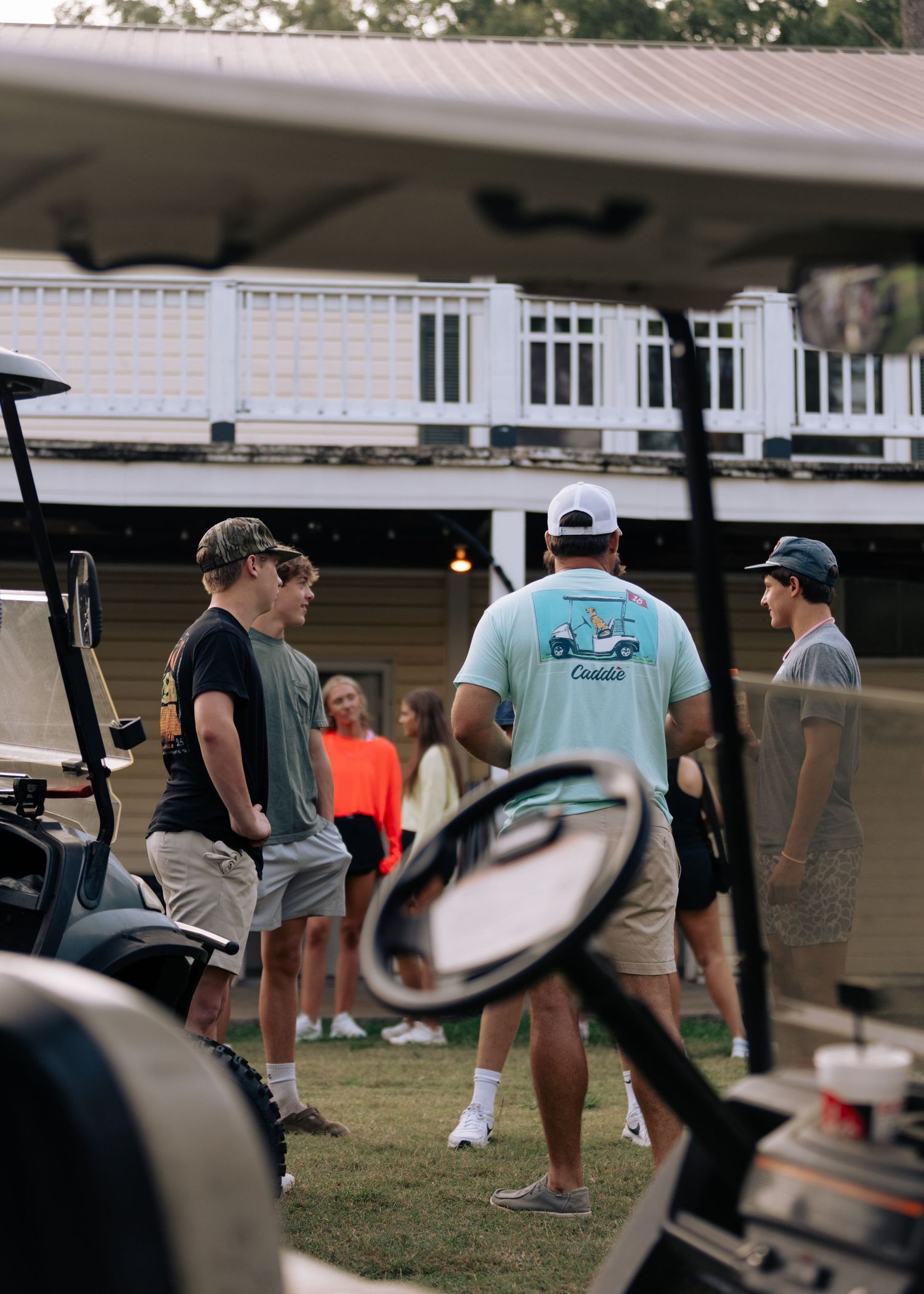 Group of young people socializing near golf carts outside a clubhouse at dusk.