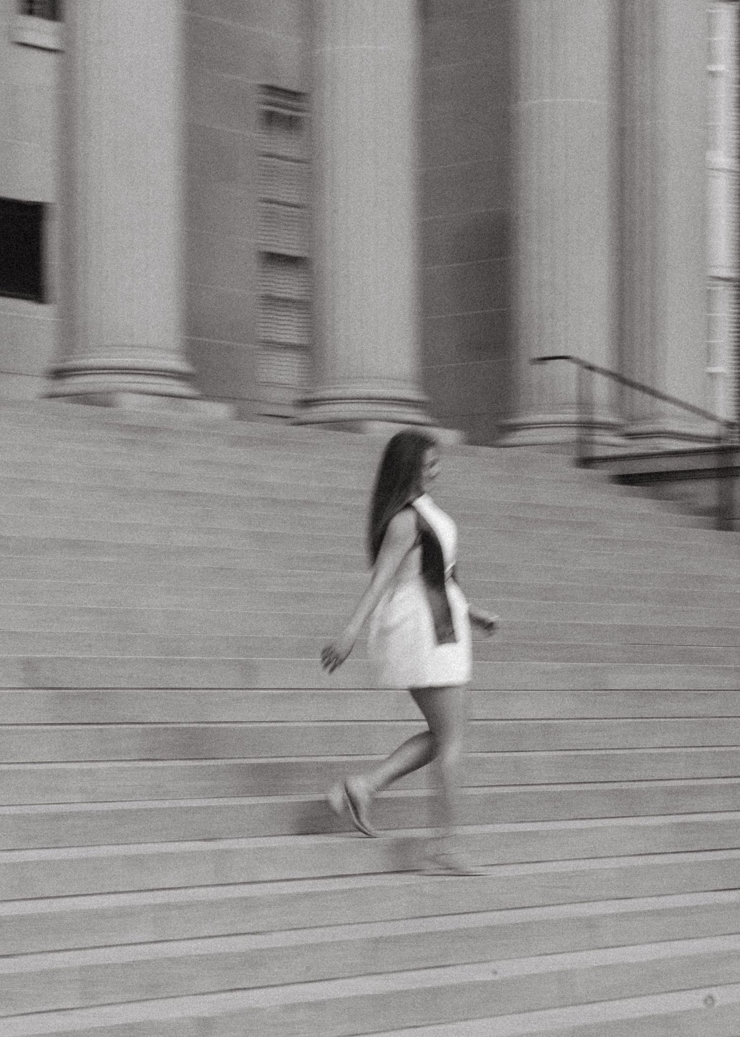 Black and white motion blur photo of graduate in white dress walking up grand stone steps.