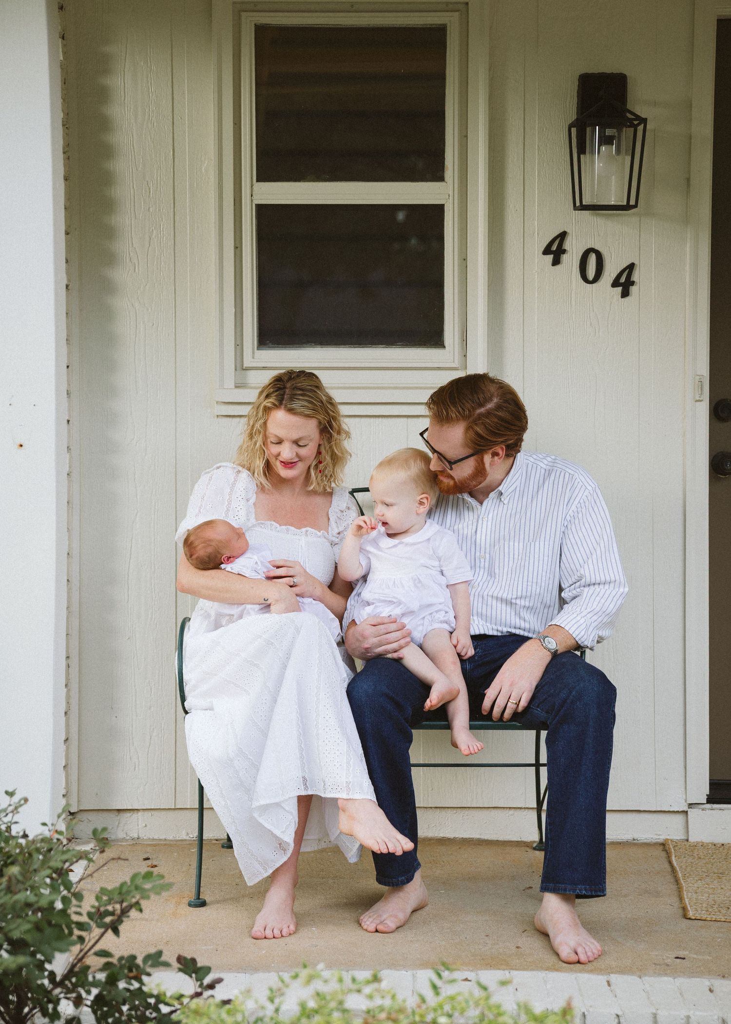A smiling couple sits barefoot on a porch bench holding a newborn baby and a curious toddler in front of a white house numbered 404.