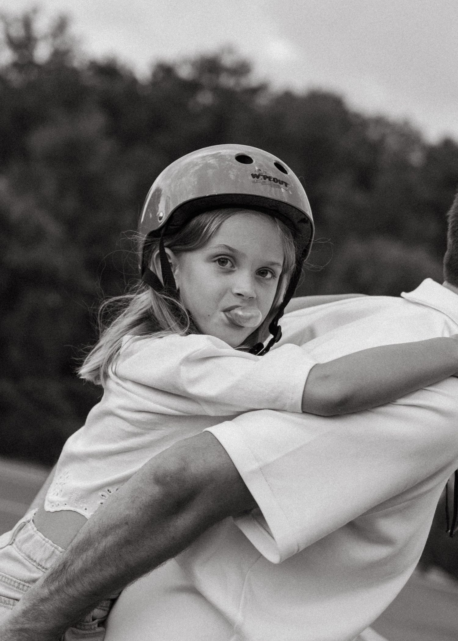 A young girl wearing a helmet sticks out her tongue in a black and white skatepark photo.