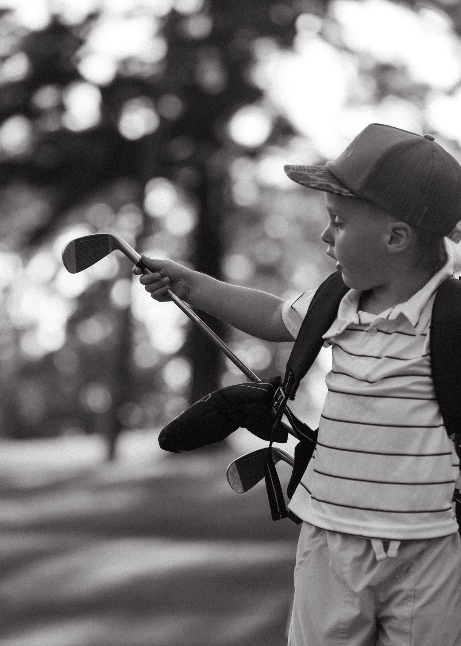 Young boy in striped shirt and cap holds golf club on course in black and white photo.