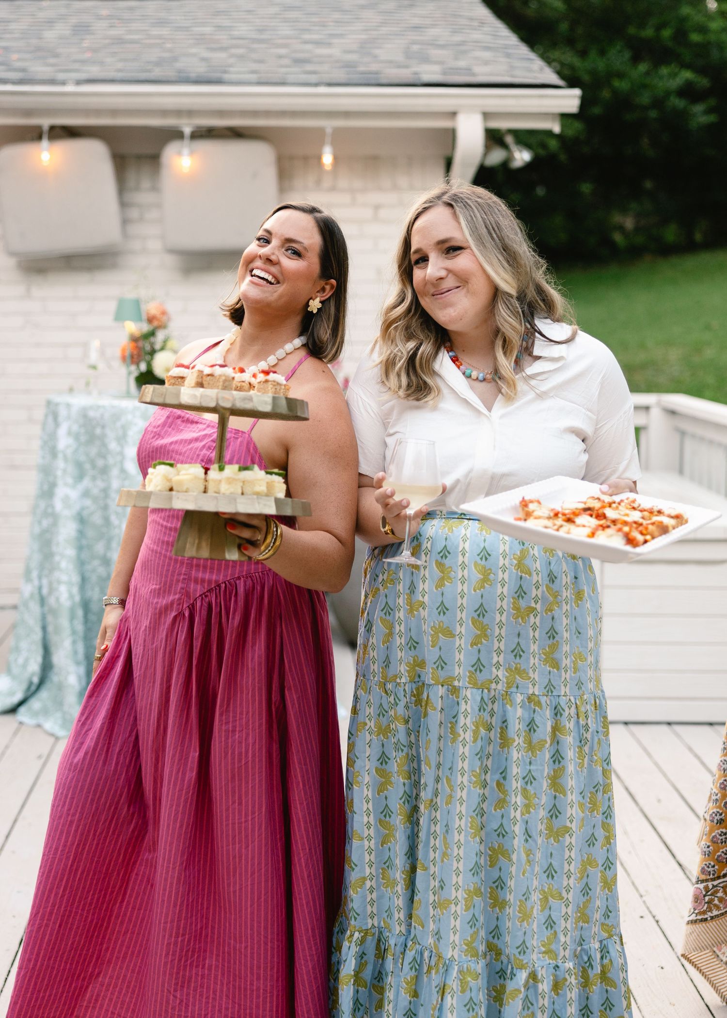 Two smiling women holding trays of desserts and appetizers at an outdoor summer garden party on a deck.